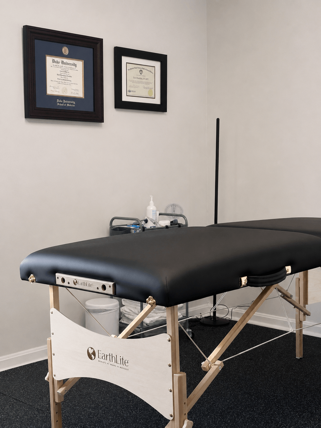 Massage table with black cover in a room with framed certificates on the wall.