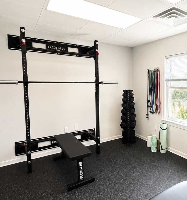 Gym room with a squat rack, bench, and resistance bands near a window, with black flooring and white walls.