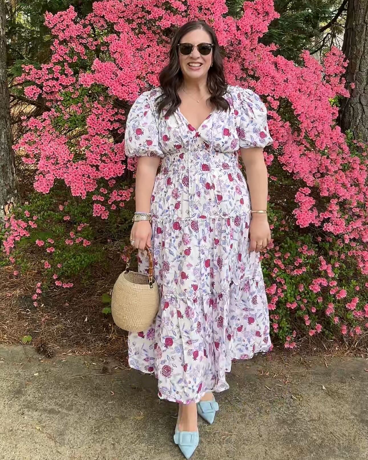 Woman in floral dress standing in front of pink flowering bush, holding a straw bag, wearing sunglasses and blue shoes.
