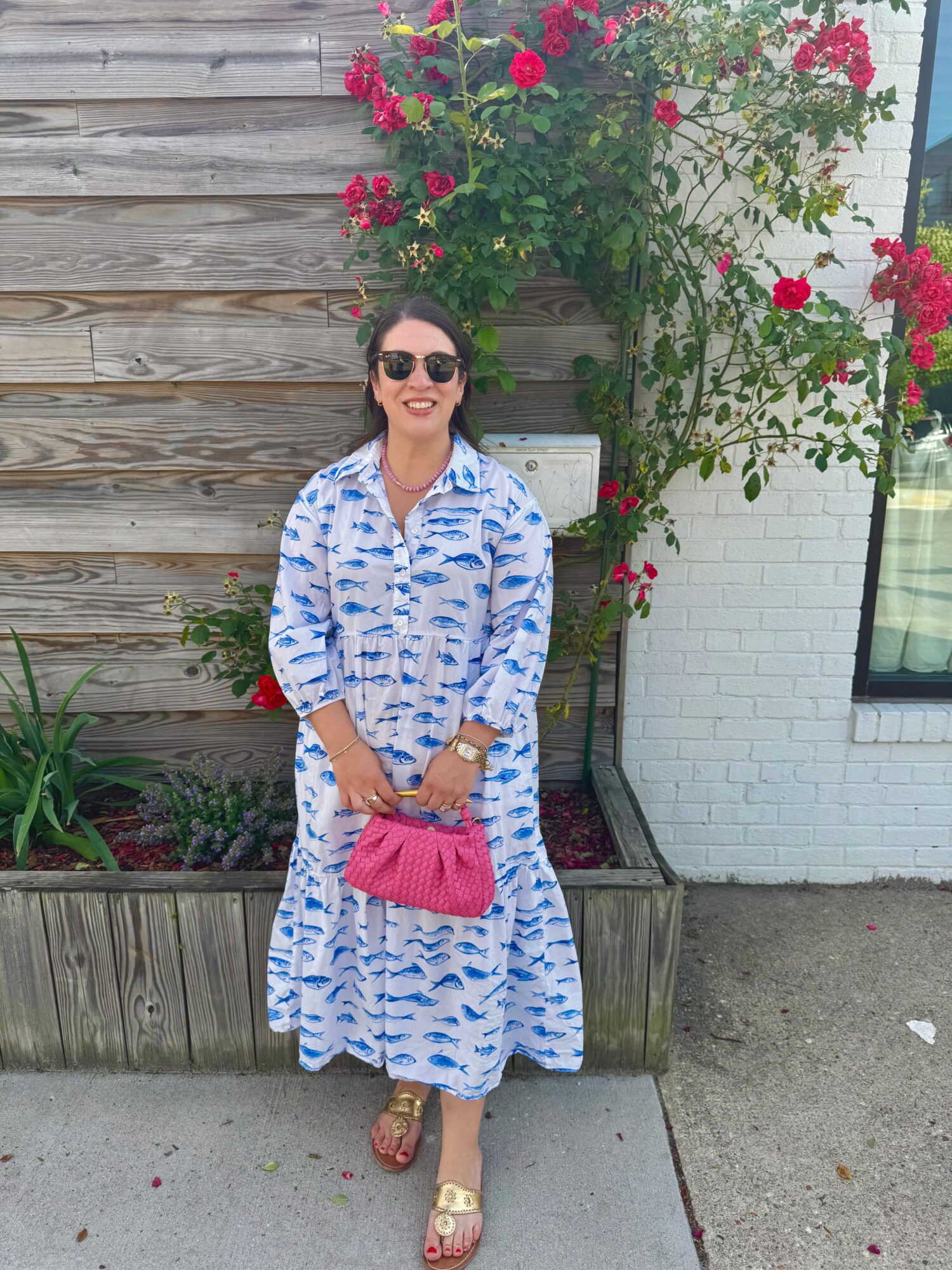 Woman in a blue and white patterned dress holding a pink purse, standing in front of a wooden fence and flowering plant, smiling, wearing sunglasses.