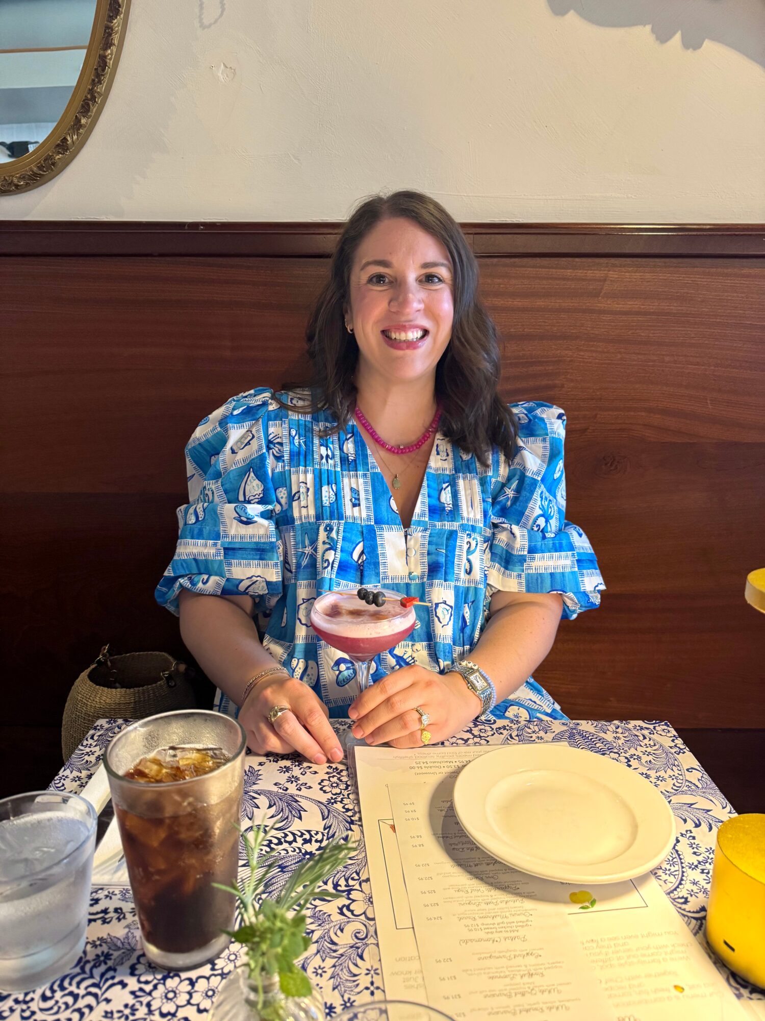 Woman with dark hair in a blue patterned dress sitting at a table, smiling, holding a dessert glass, in a restaurant.
