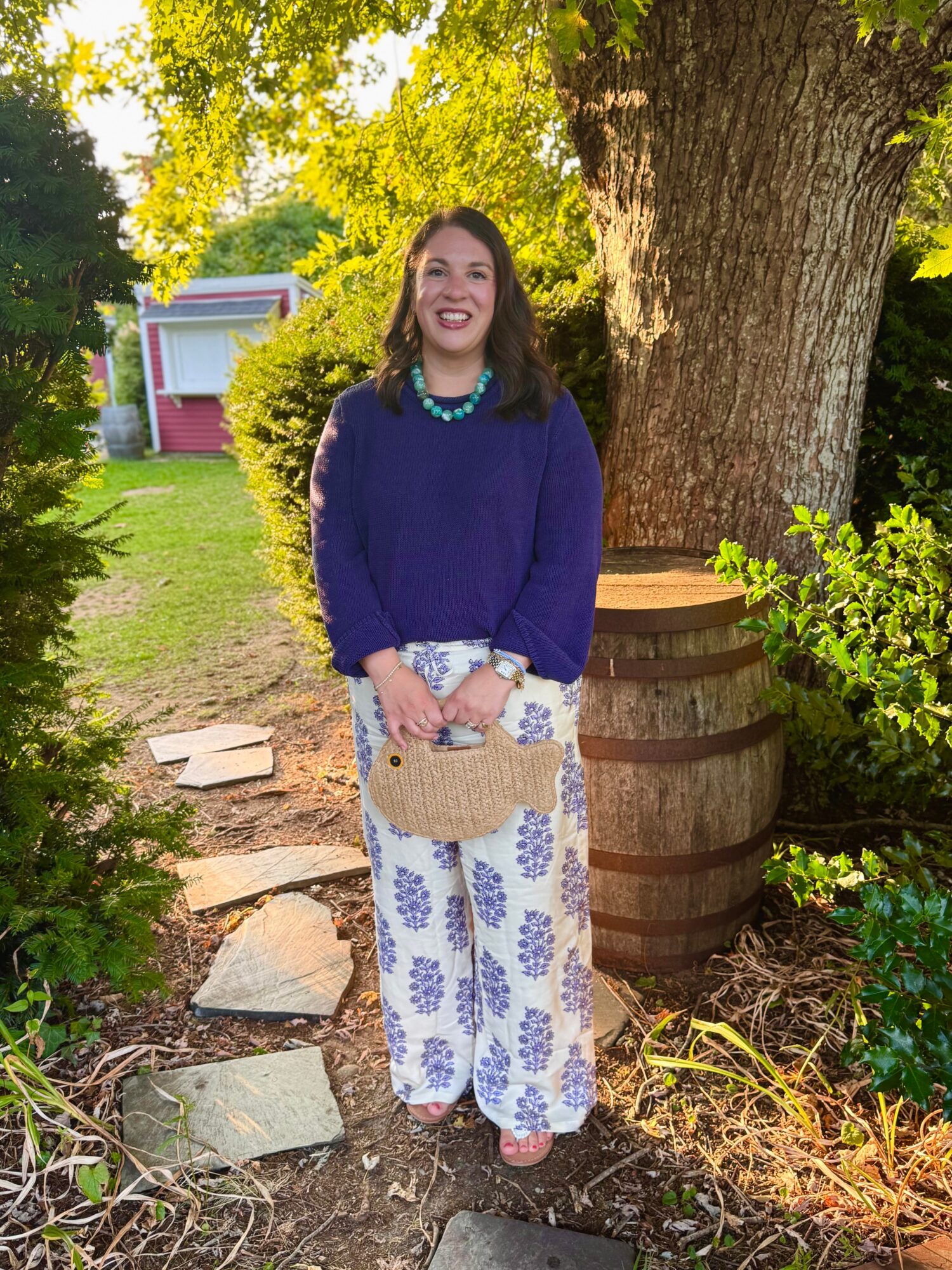 Woman standing outdoors near a tree, smiling, holding a woven bag, wearing a blue sweater and patterned pants.