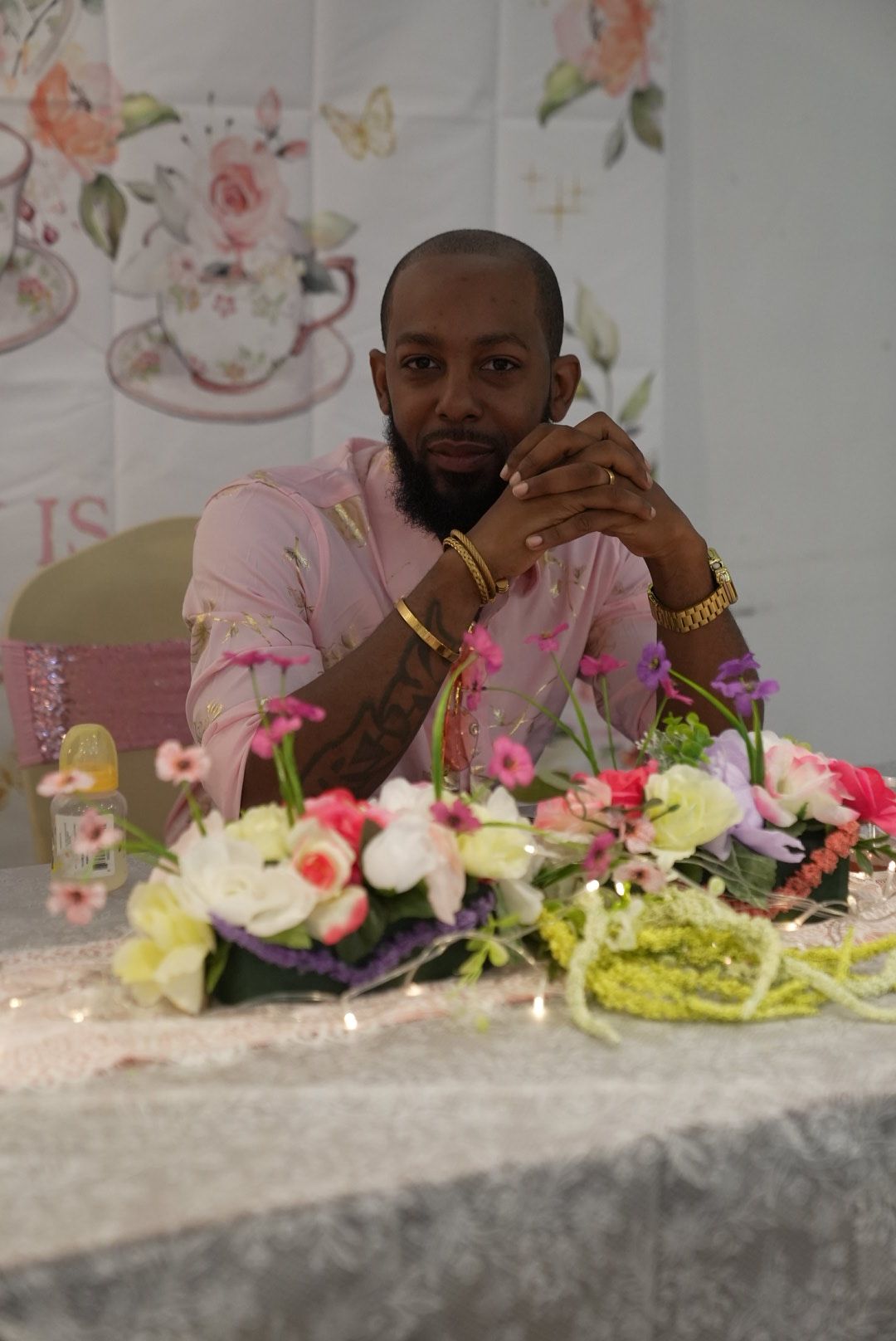 Woman in pink dress sitting at table with floral decorations, holding a microphone, in a decorated event space.