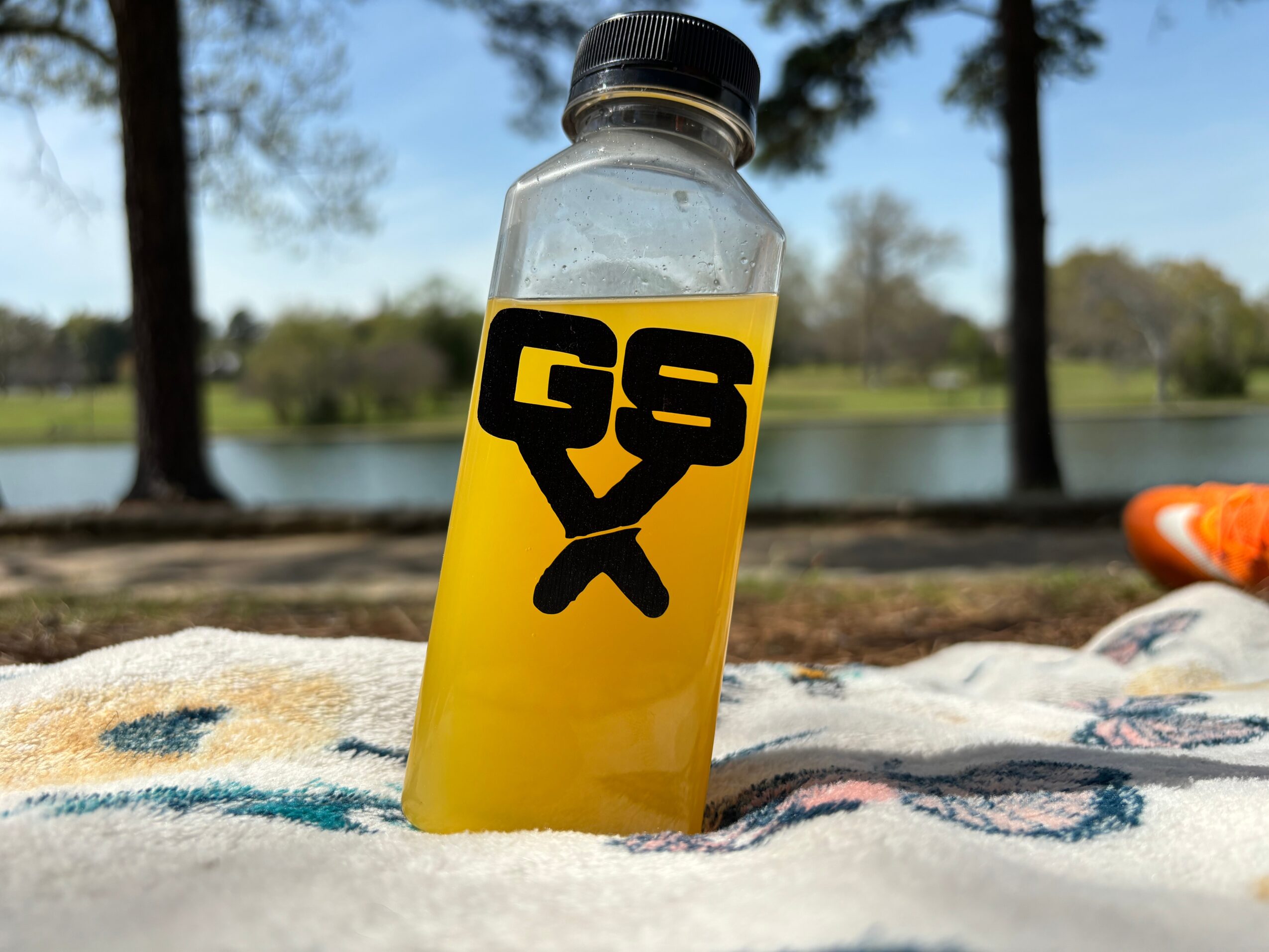 A clear plastic bottle with a black cap and yellow label, partially buried in sand outdoors with trees and water in background.
