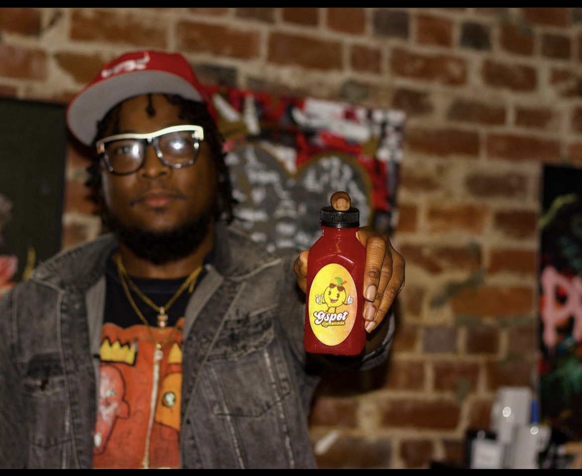Person with glasses and a red cap holding a red bottle with a yellow label, standing in front of a brick wall.