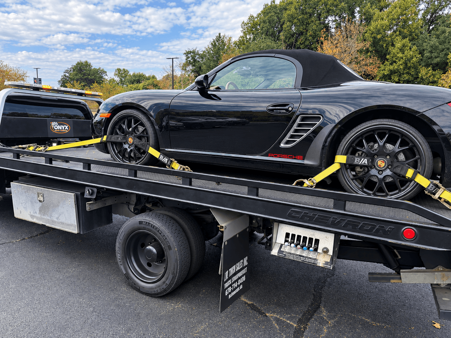 Black convertible sports car on a flatbed tow truck, with trees and a partly cloudy sky in the background.