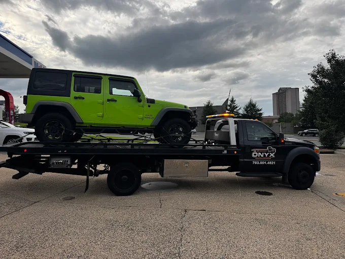 Flatbed tow truck carrying a green Jeep with a black roof, parked on a street with cloudy sky and trees in background.