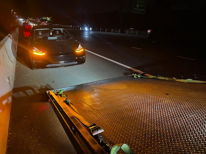 Nighttime scene showing a black sports car on a highway with other vehicles, and a flatbed truck in the foreground.