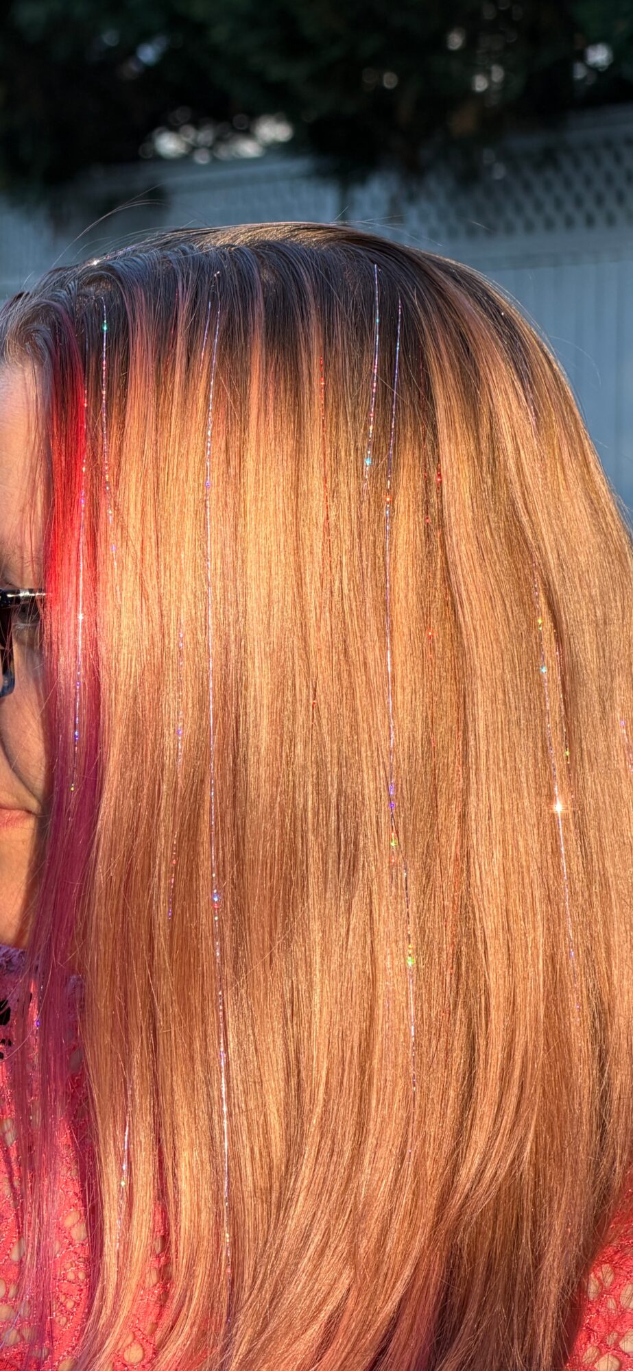 Close-up of a person's hair with blonde and pink streaks, outdoors with a fence and trees in background.