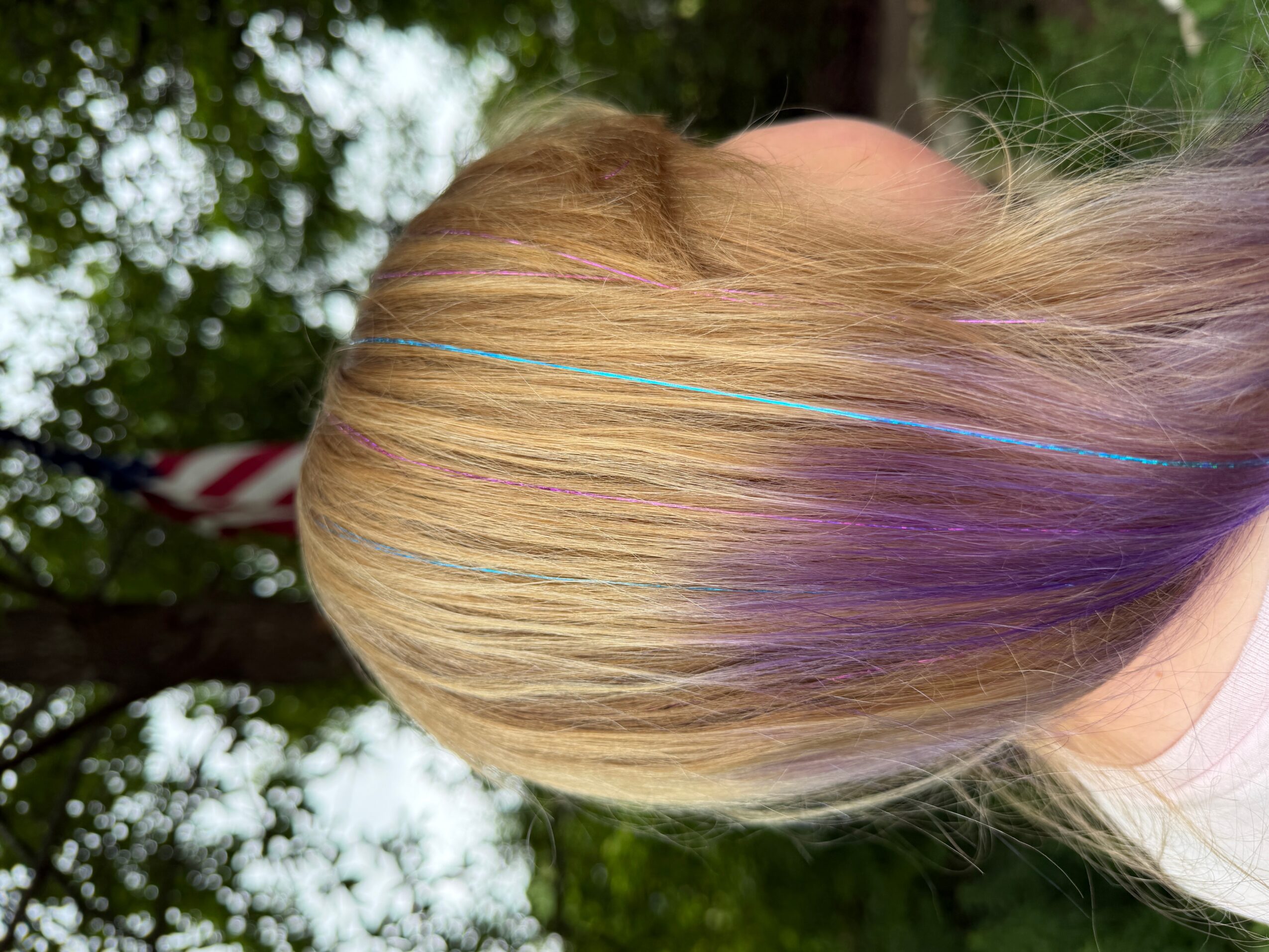 Side view of a person with blonde hair featuring purple streaks, outdoors with trees in the background.