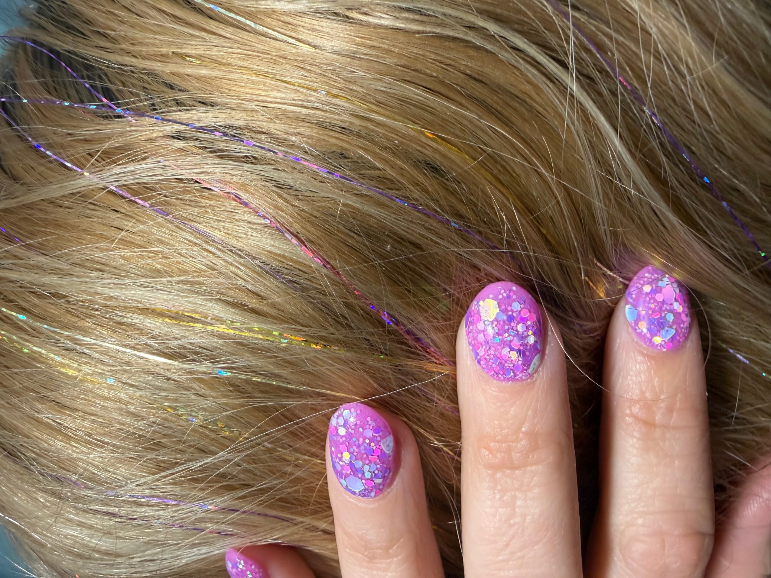 Close-up of fingers with purple glittery nail polish, hair with colorful streaks, and a portion of face visible.