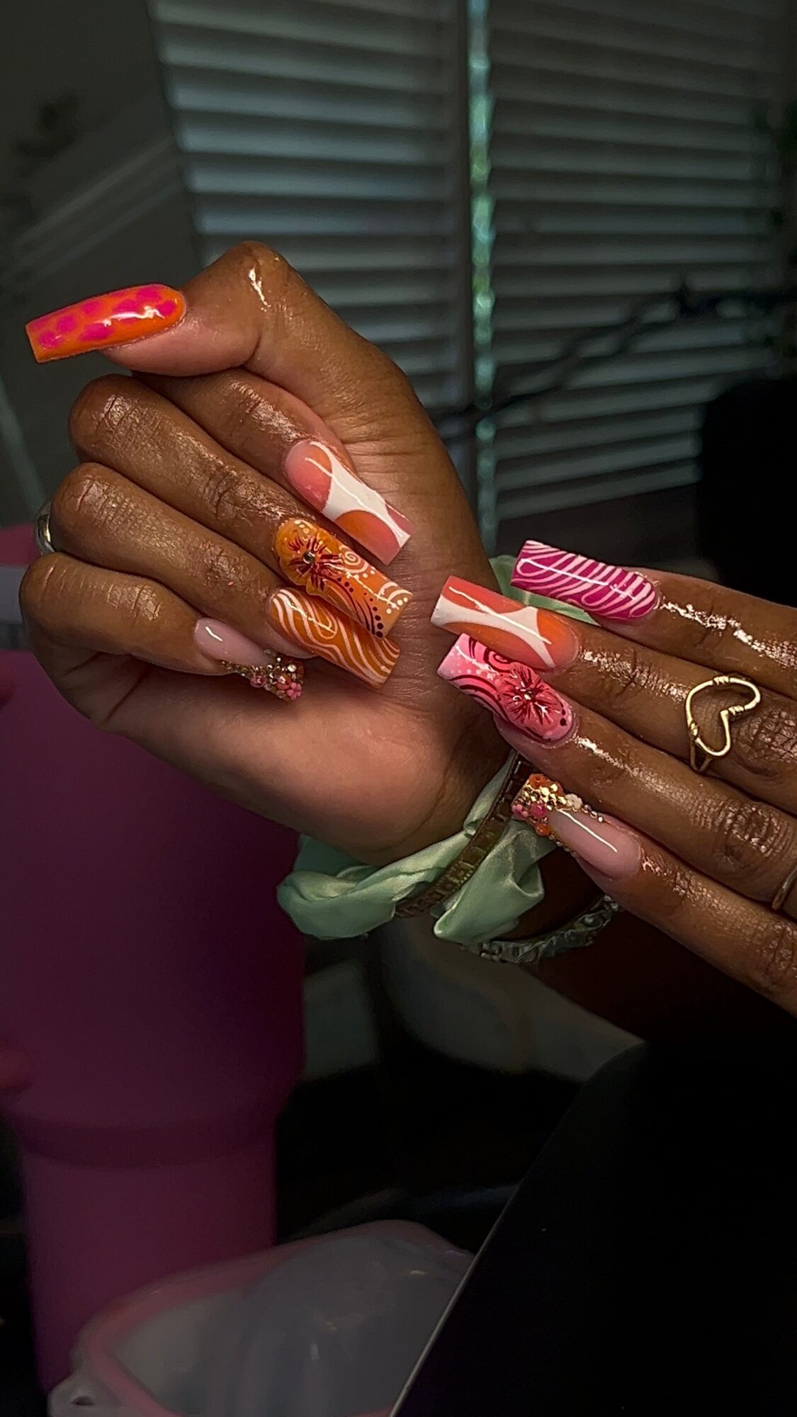 Close-up of hands with colorful, decorated nails, including pink, orange, and gold designs, and a heart-shaped ring.