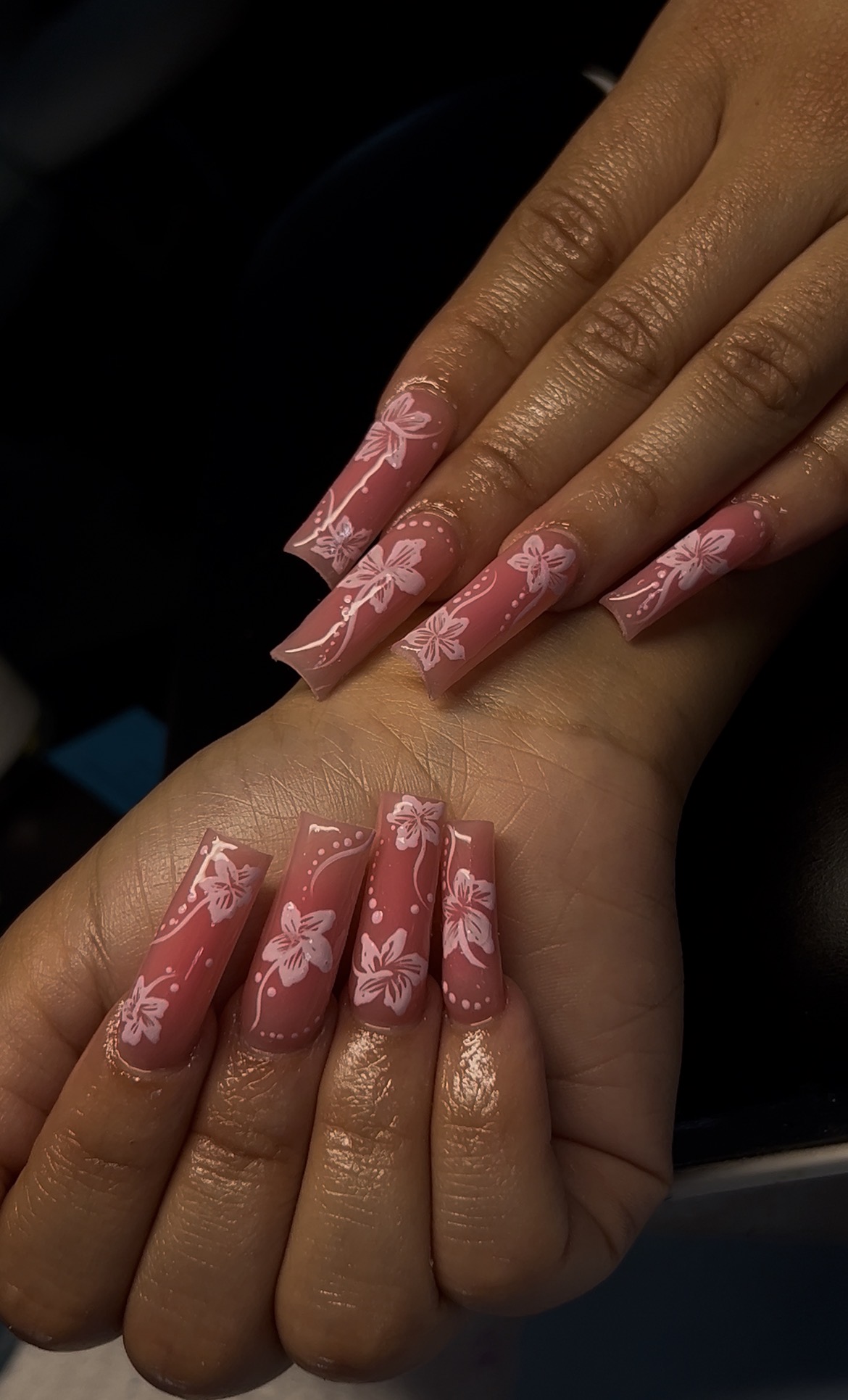 Two hands with long, decorated pink nails featuring white floral designs, positioned one above the other.