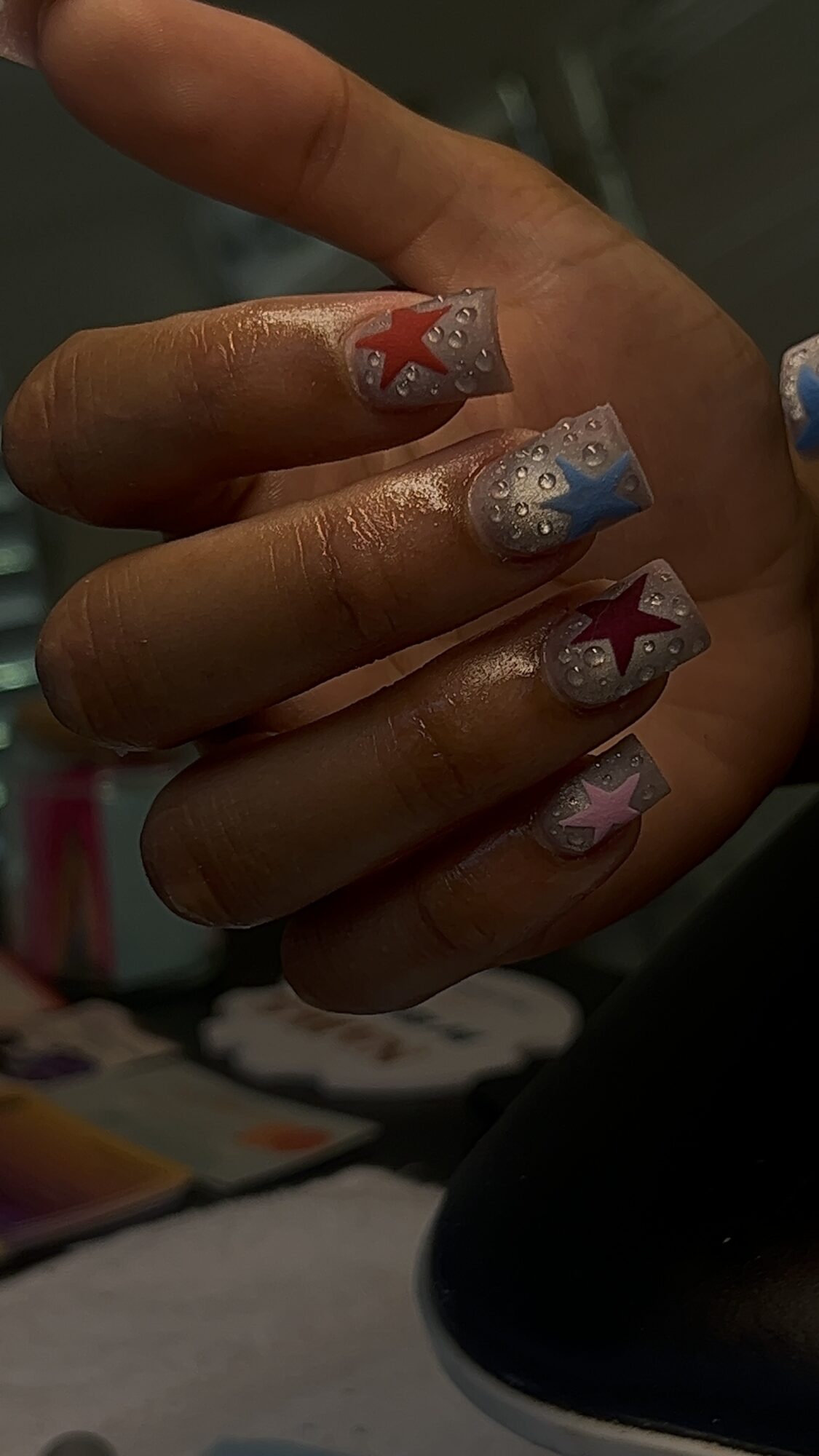 Hand with decorated nails featuring red, blue, and silver star designs, some with glitter, against a dark background.