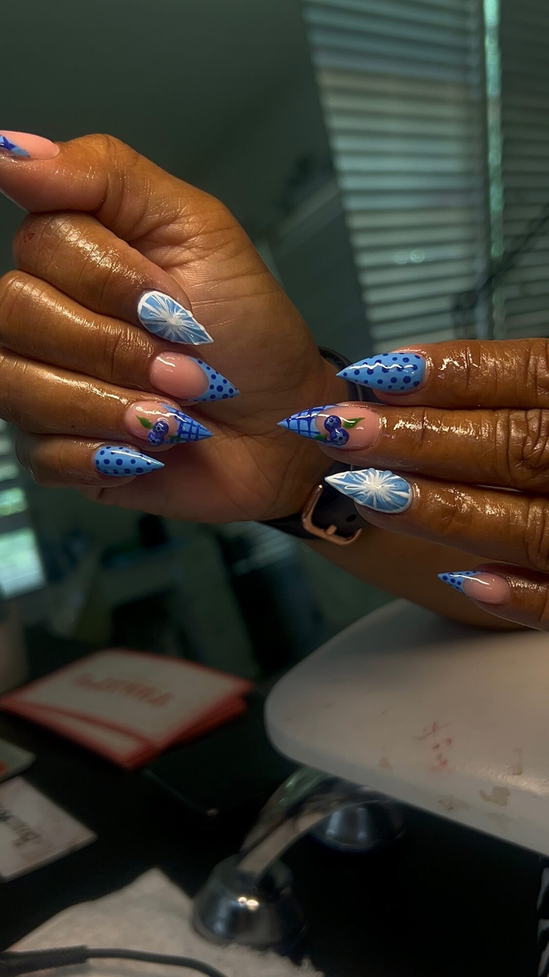 Two hands with blue and white patterned nails, one hand resting on the other, indoors with blinds in background.