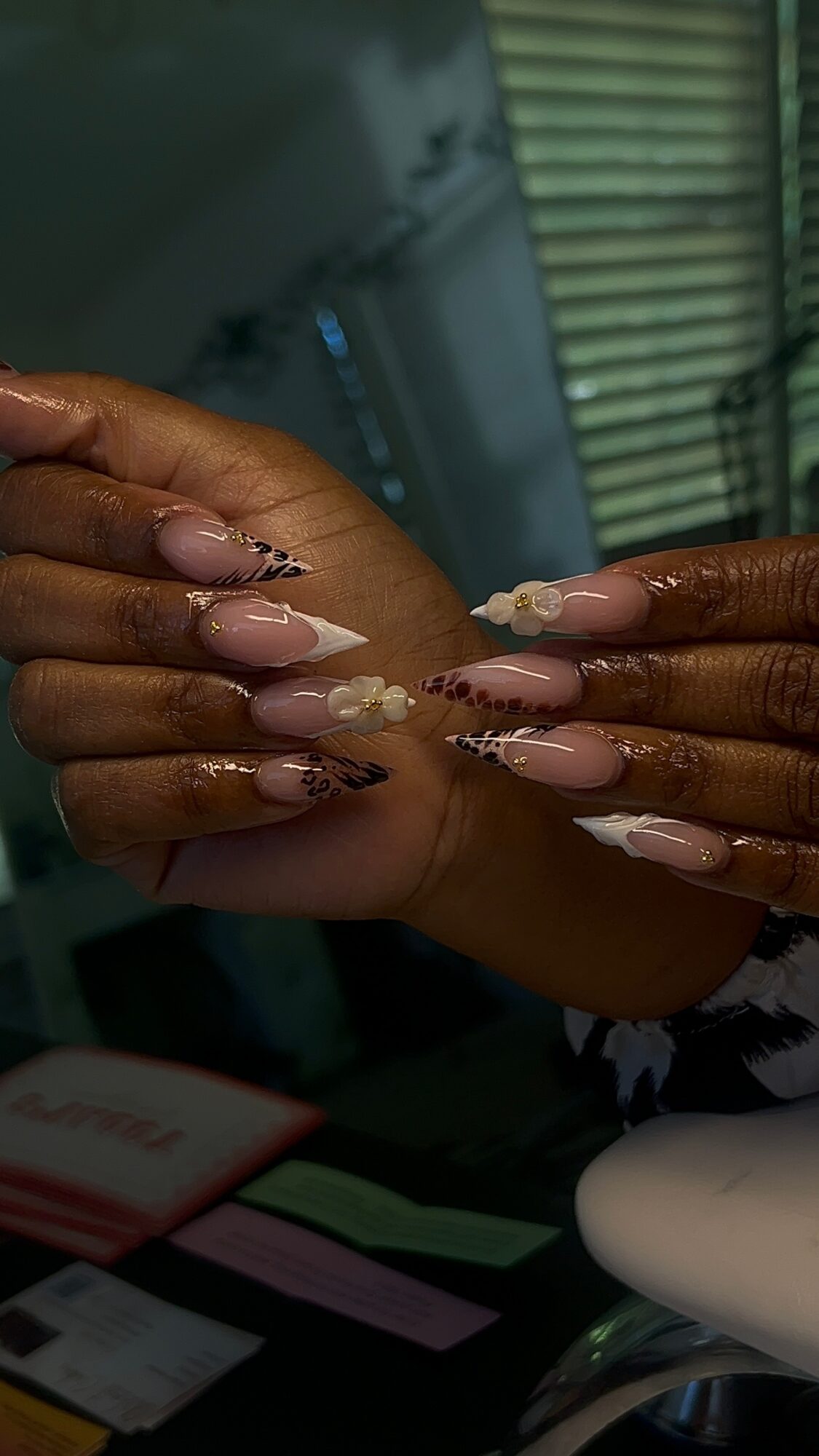 Two hands with long, decorated nails, holding each other, in a setting with a window and various objects in the background.