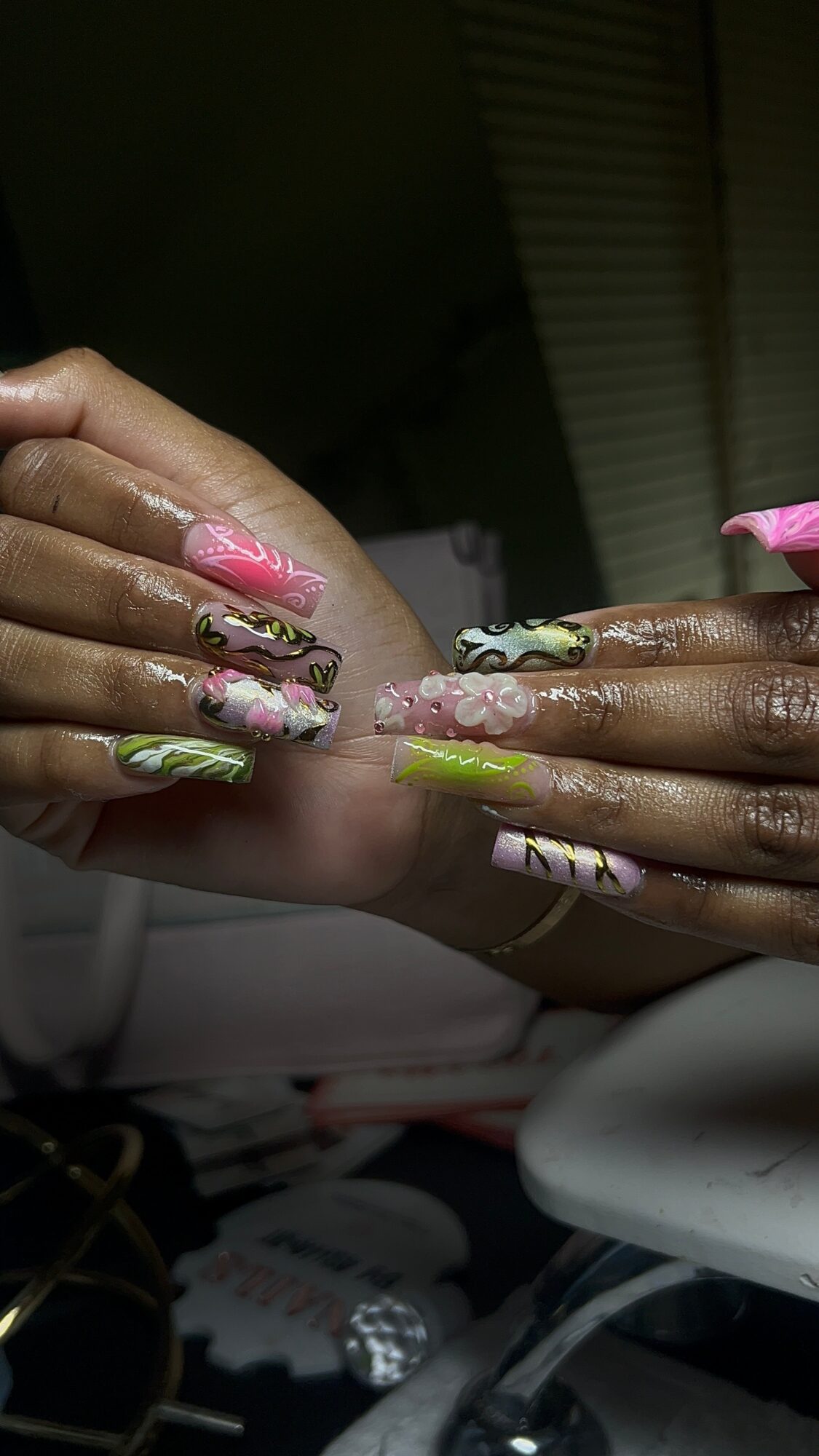 Two hands with colorful, decorated nails resting on a surface, with a dark background and window blinds visible.
