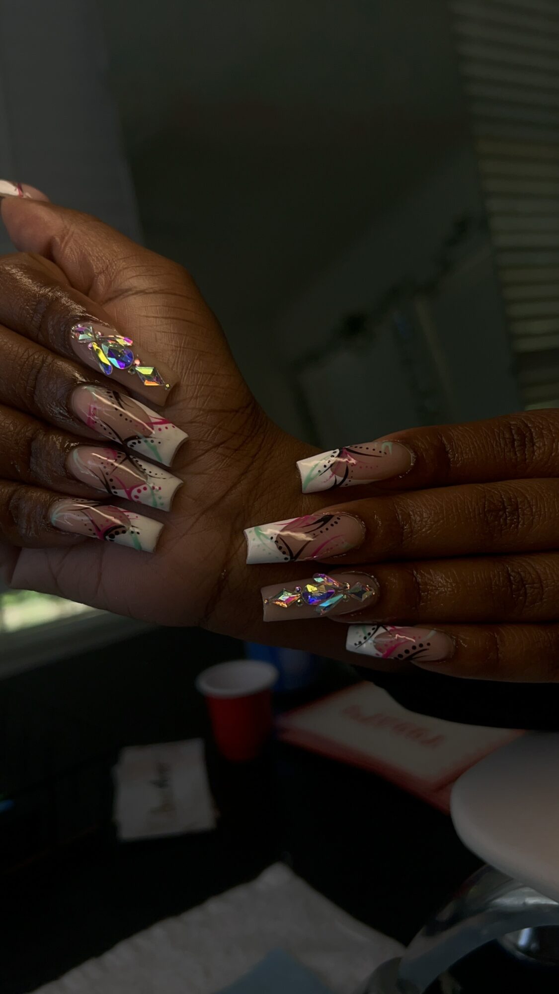 Close-up of hands with long, decorated nails, some with holographic designs, in a room with a dark background.