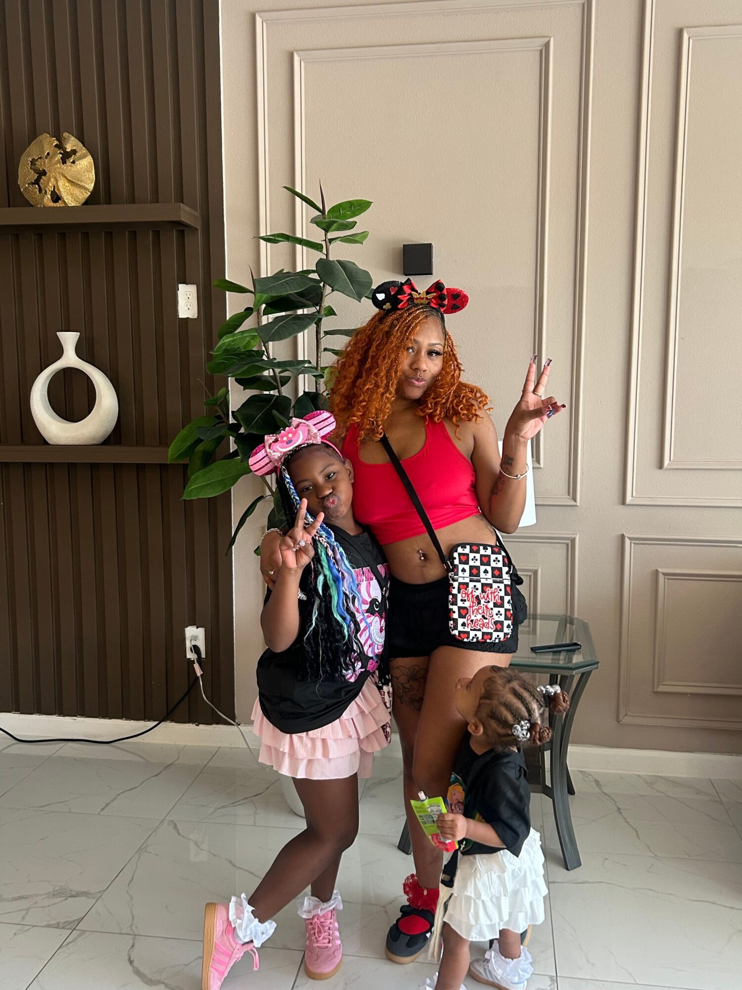 Two young girls standing indoors, one making a peace sign, with a plant and decorative wall behind them.