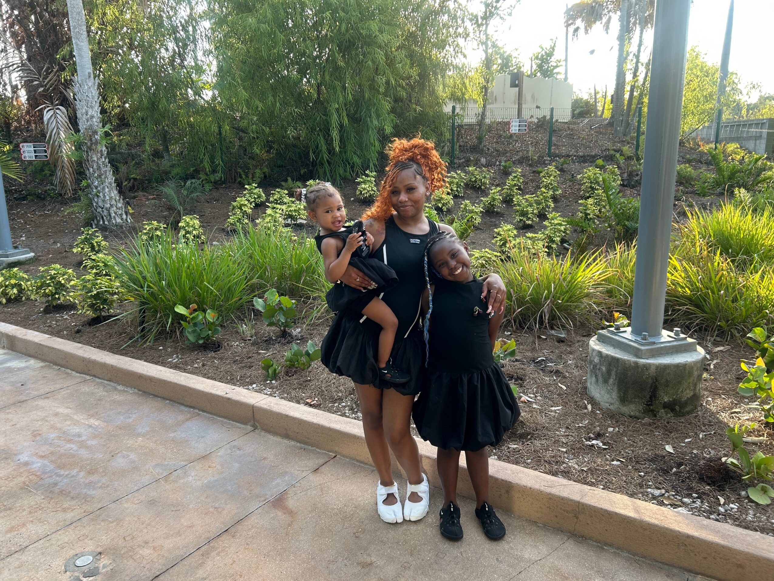 Woman with two children standing outdoors on a sidewalk, surrounded by greenery and plants.