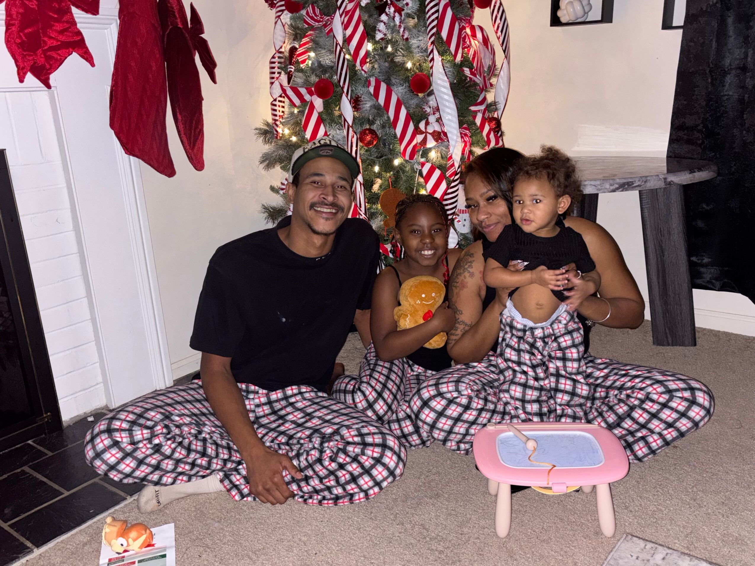 Four people sitting on the floor in front of a decorated Christmas tree, holding a small orange pumpkin, with a pink table nearby.