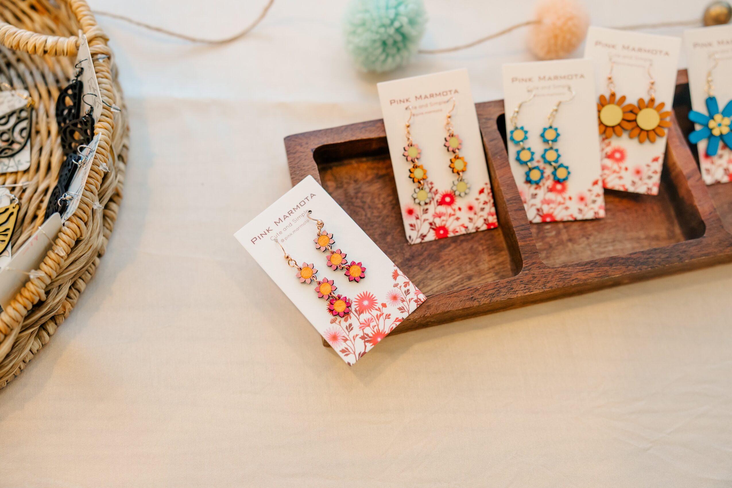 Display of colorful earrings on cards, arranged on a wooden tray, with a basket and fuzzy pom-poms nearby, on a light surface.