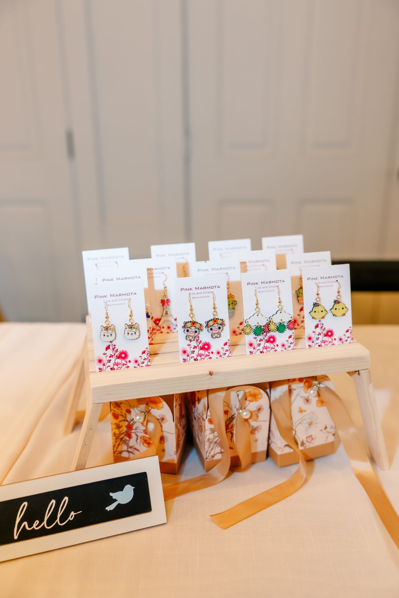 Display of jewelry cards on a wooden stand with small bags below, in front of a white door, with a 'hello' sign.