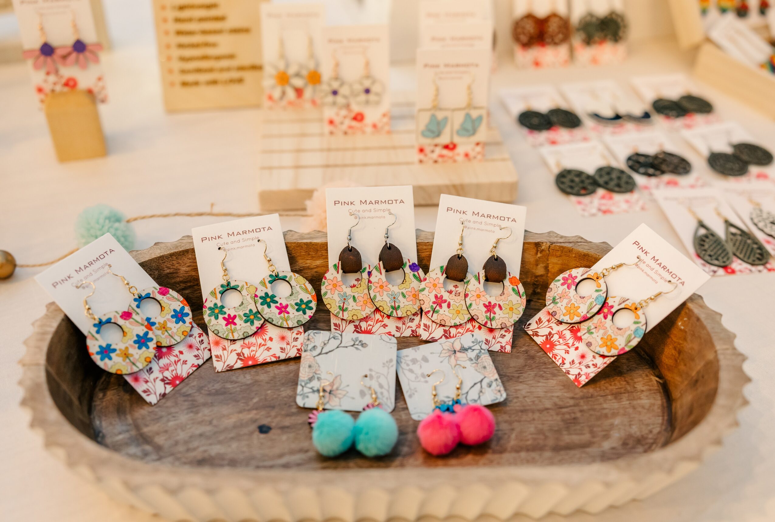 Display of colorful earrings and accessories on a wooden tray with jewelry in the background.