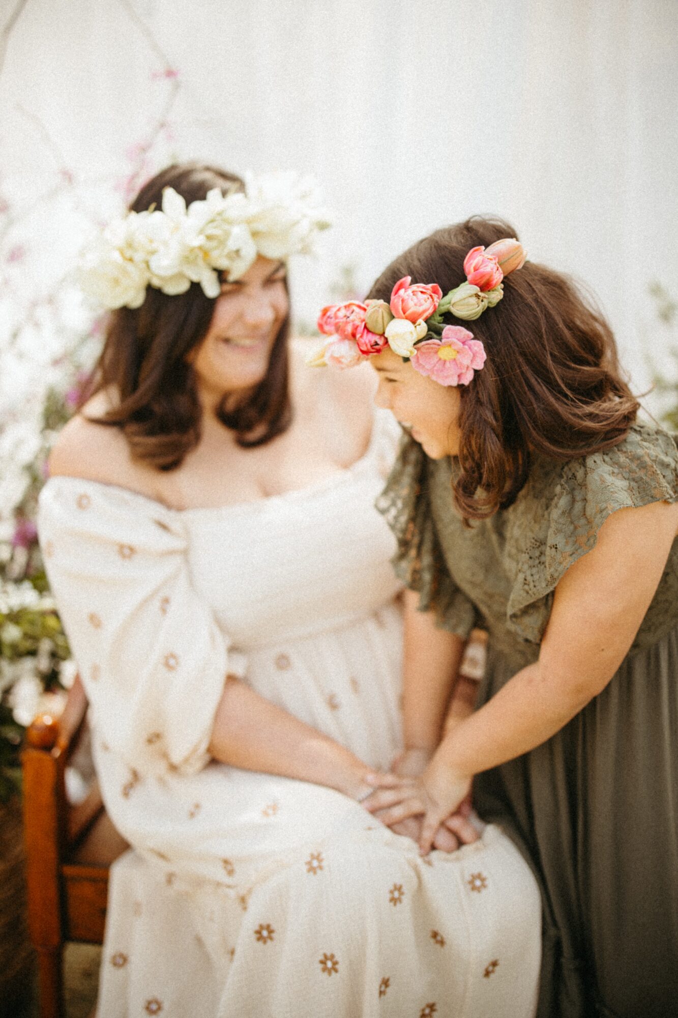 Two women wearing flower crowns, one sitting and one leaning in, holding hands, in a bright setting.