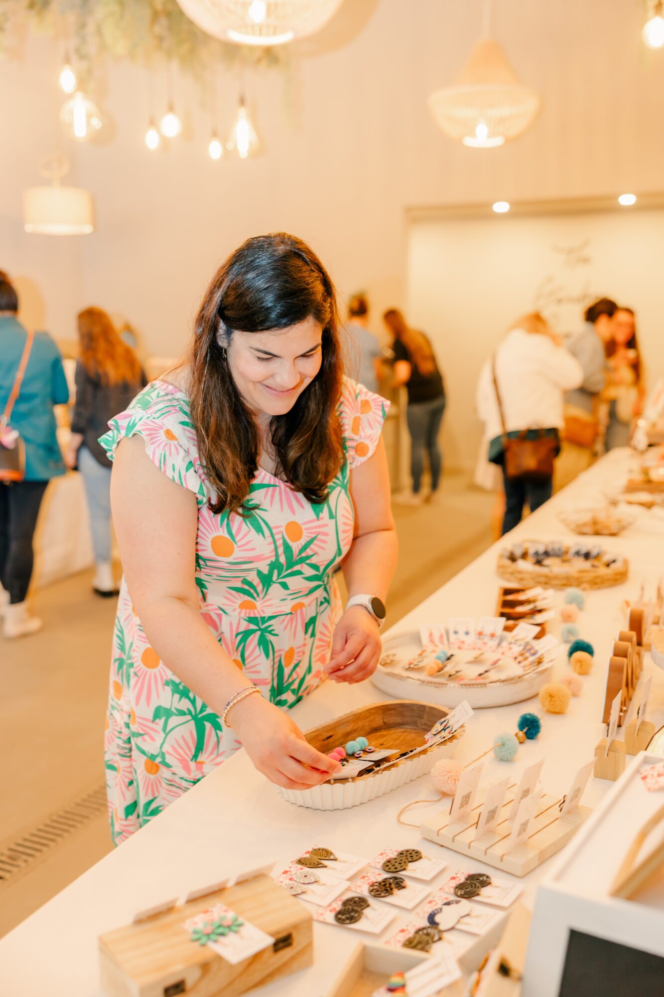 Woman in a colorful dress browsing items on a table at an indoor event, with people in the background.