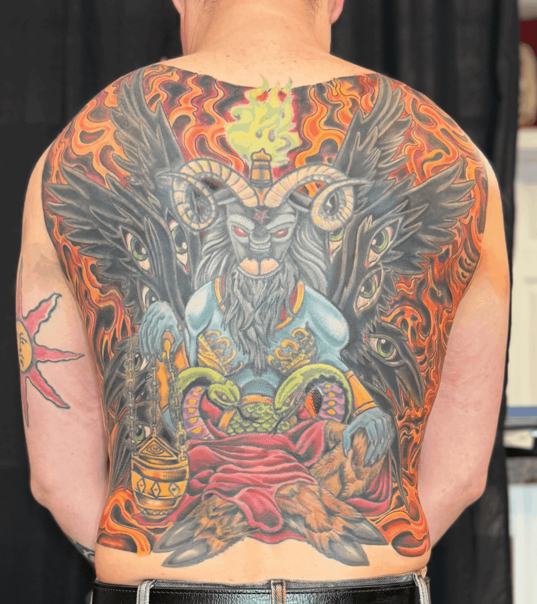Colorful tattoo of a goat with large horns, surrounded by flames and two black wolves, covering the person's back.