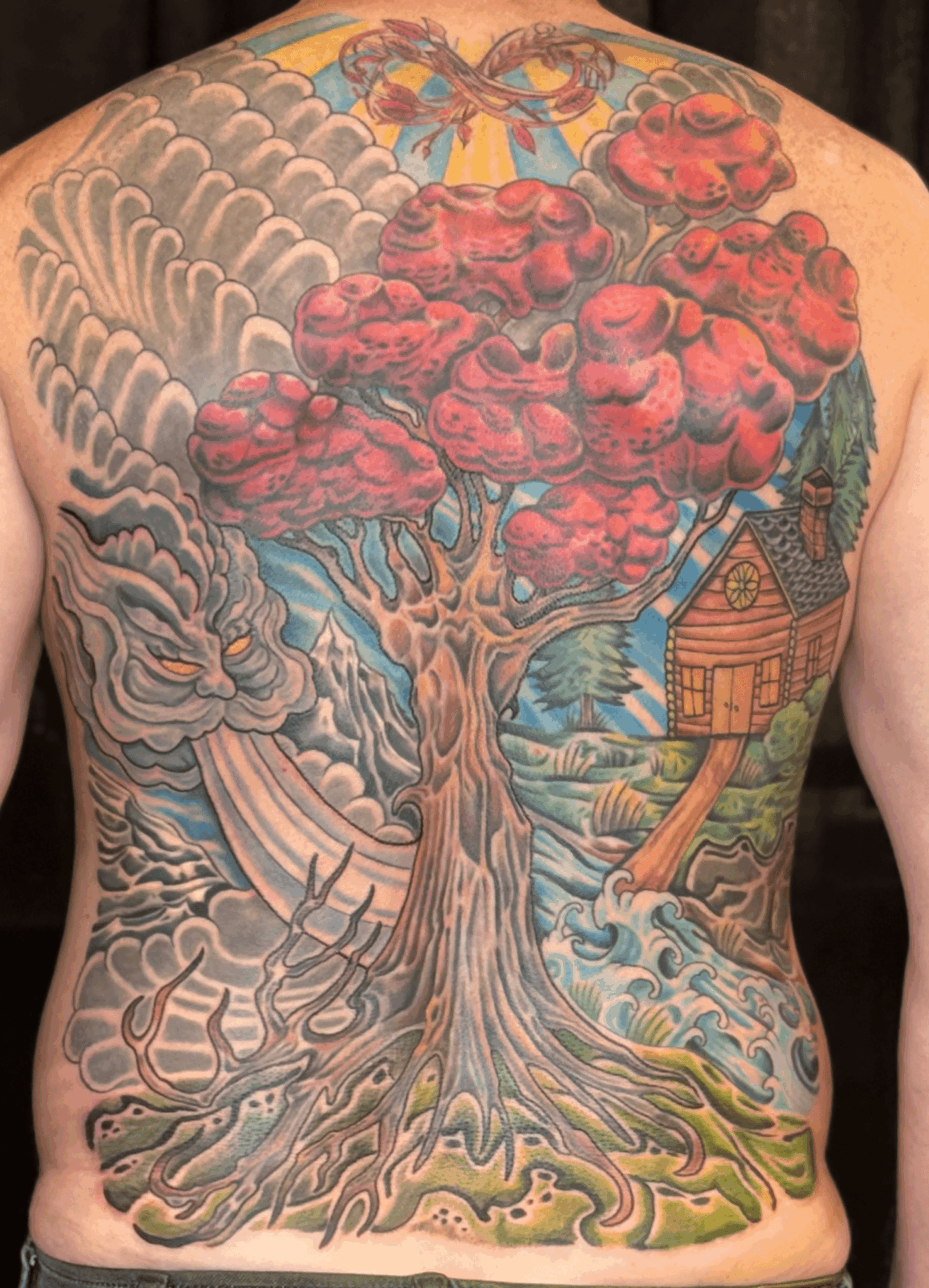 Colorful tattoo of a large tree with red foliage, a house, and swirling clouds on a person's back.