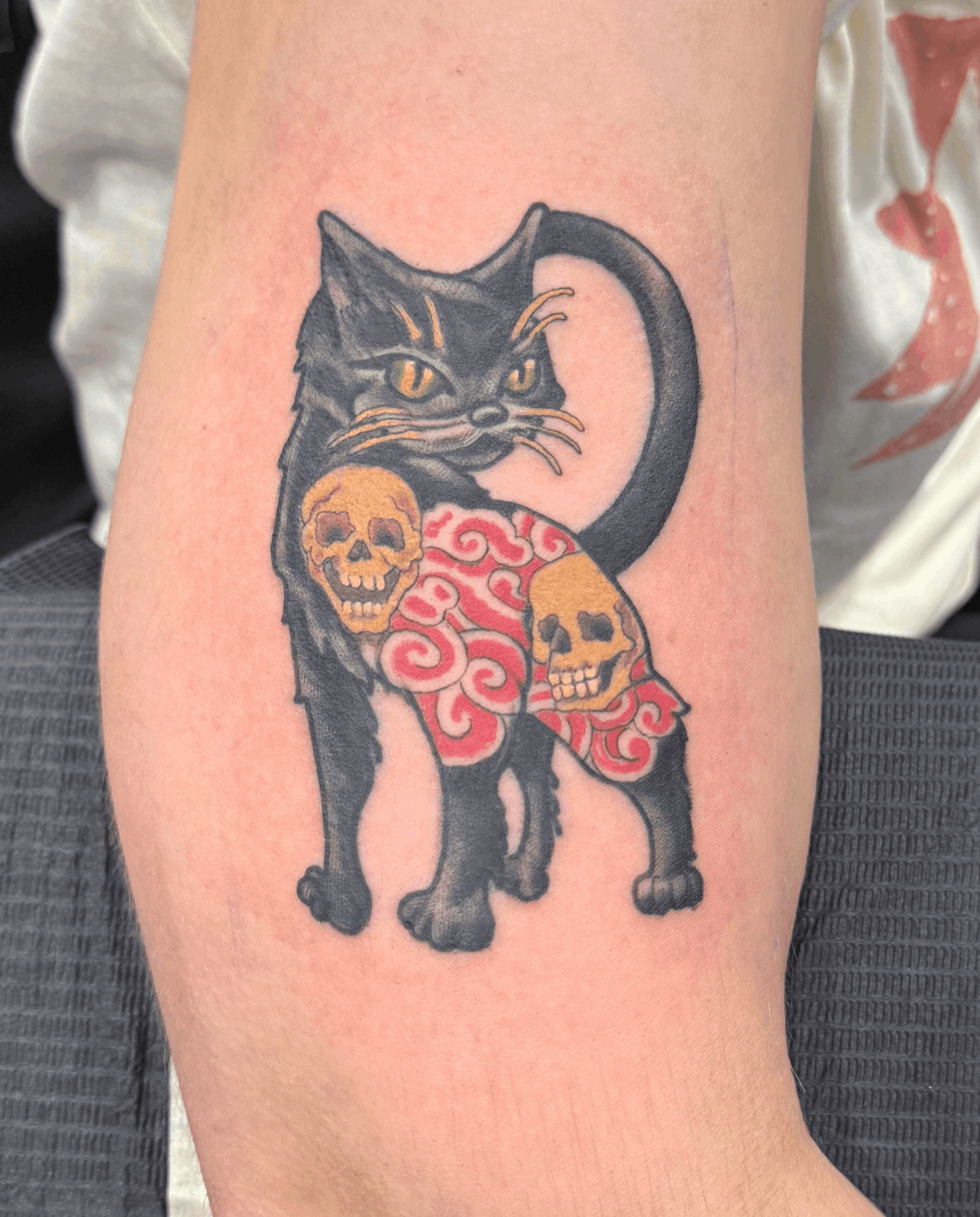 Tattoo of a black cat with yellow eyes, decorated with pink and red swirls, and skulls on its body.