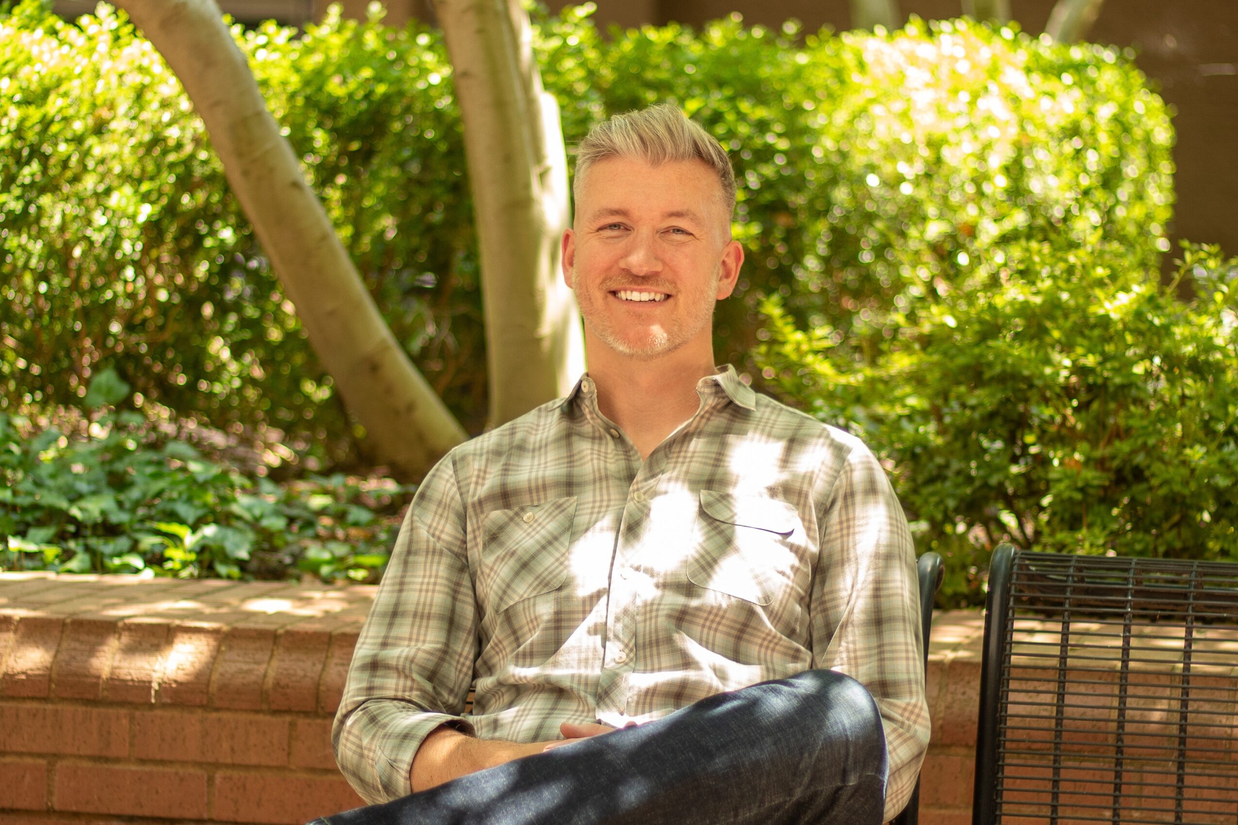 Smiling man with gray hair in a plaid shirt sitting outdoors with greenery and a brick wall behind him.