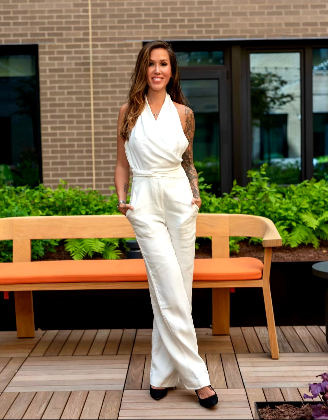 Woman in white jumpsuit standing outdoors in front of a bench and greenery, smiling, with a brick building behind.