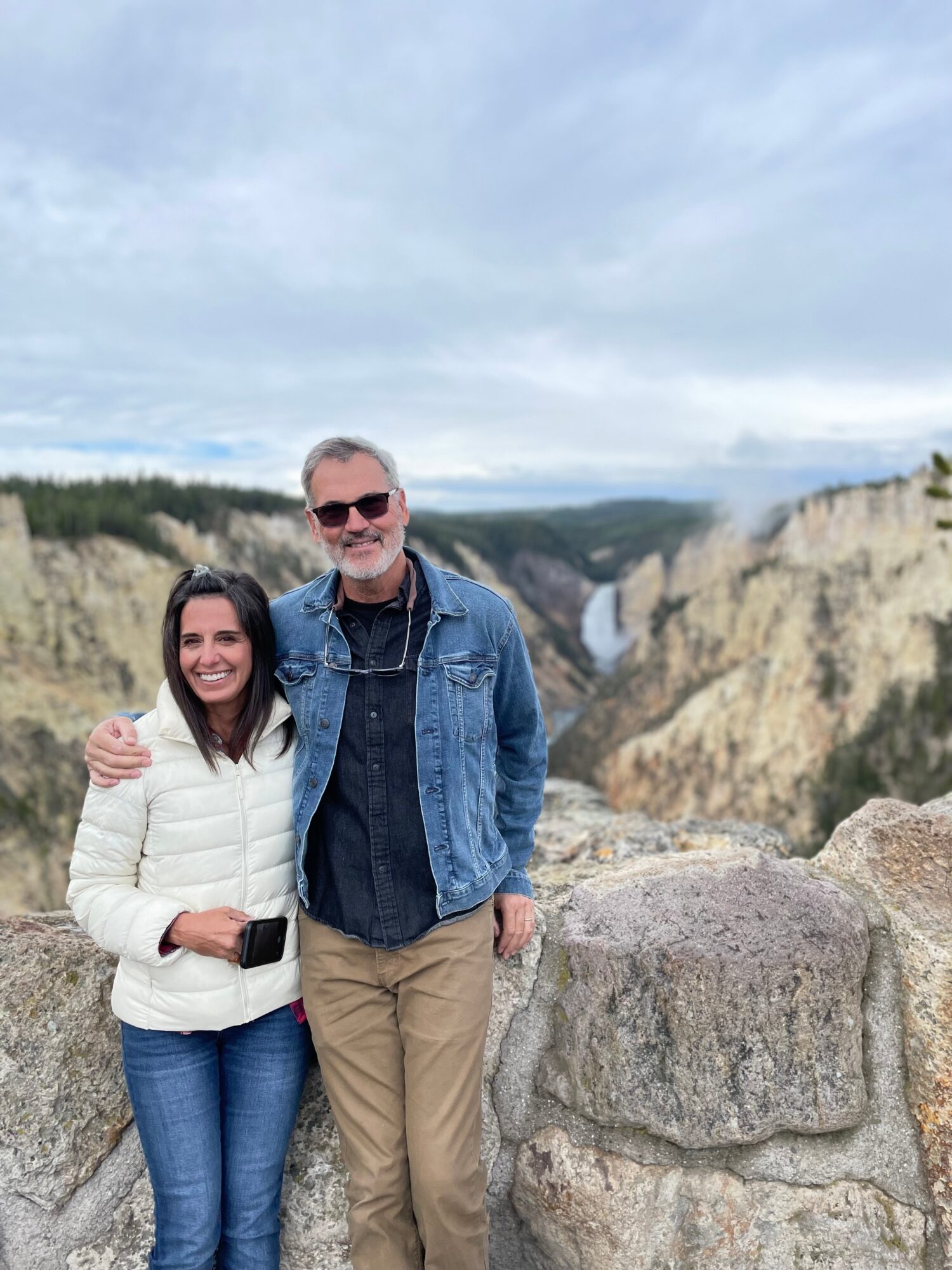 Two people stand outdoors near a rocky ledge with a canyon and waterfall in the background, smiling at the camera.