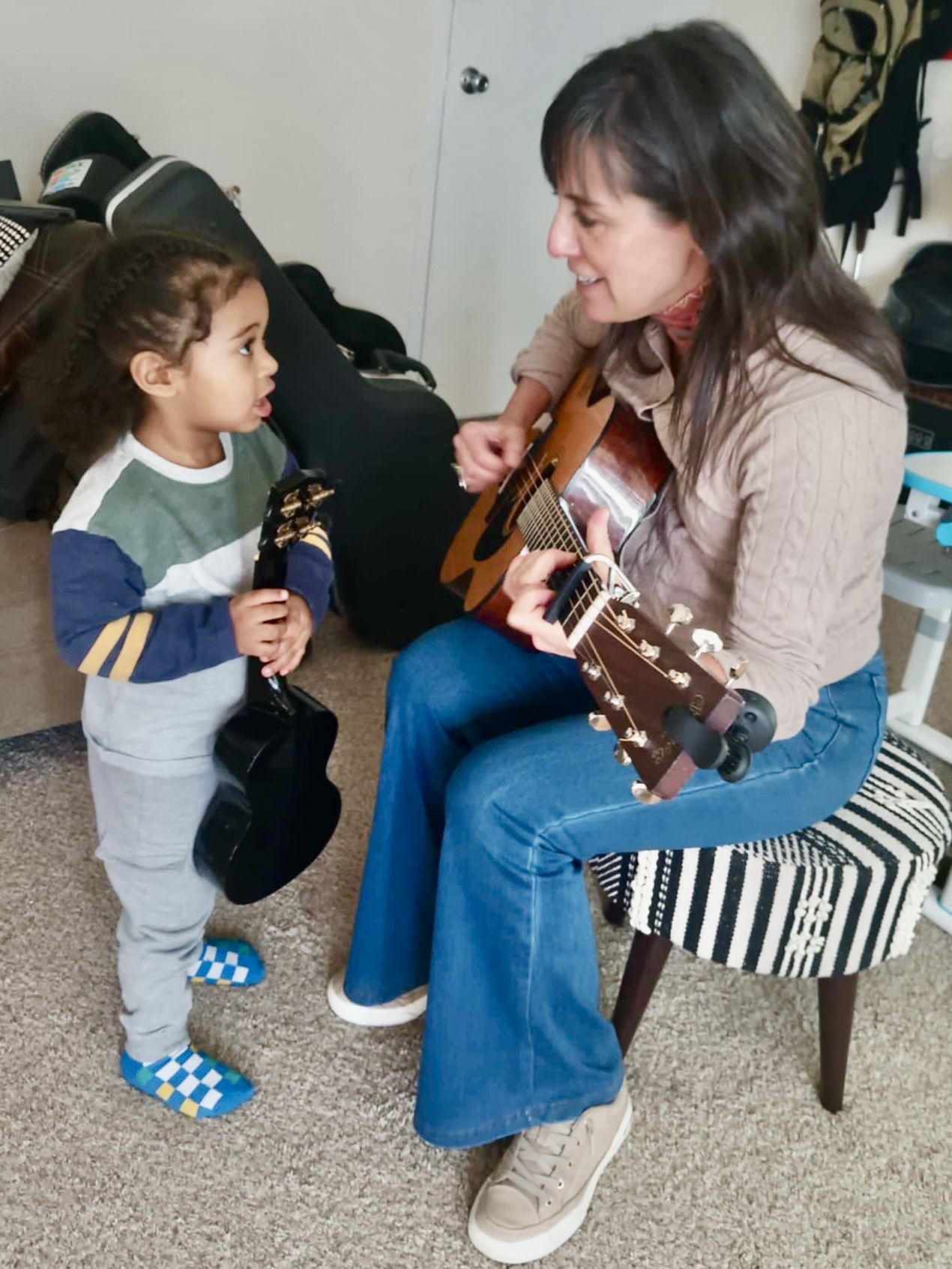 Adult woman playing guitar while talking to a young girl indoors, with a striped chair and clothing in the background.
