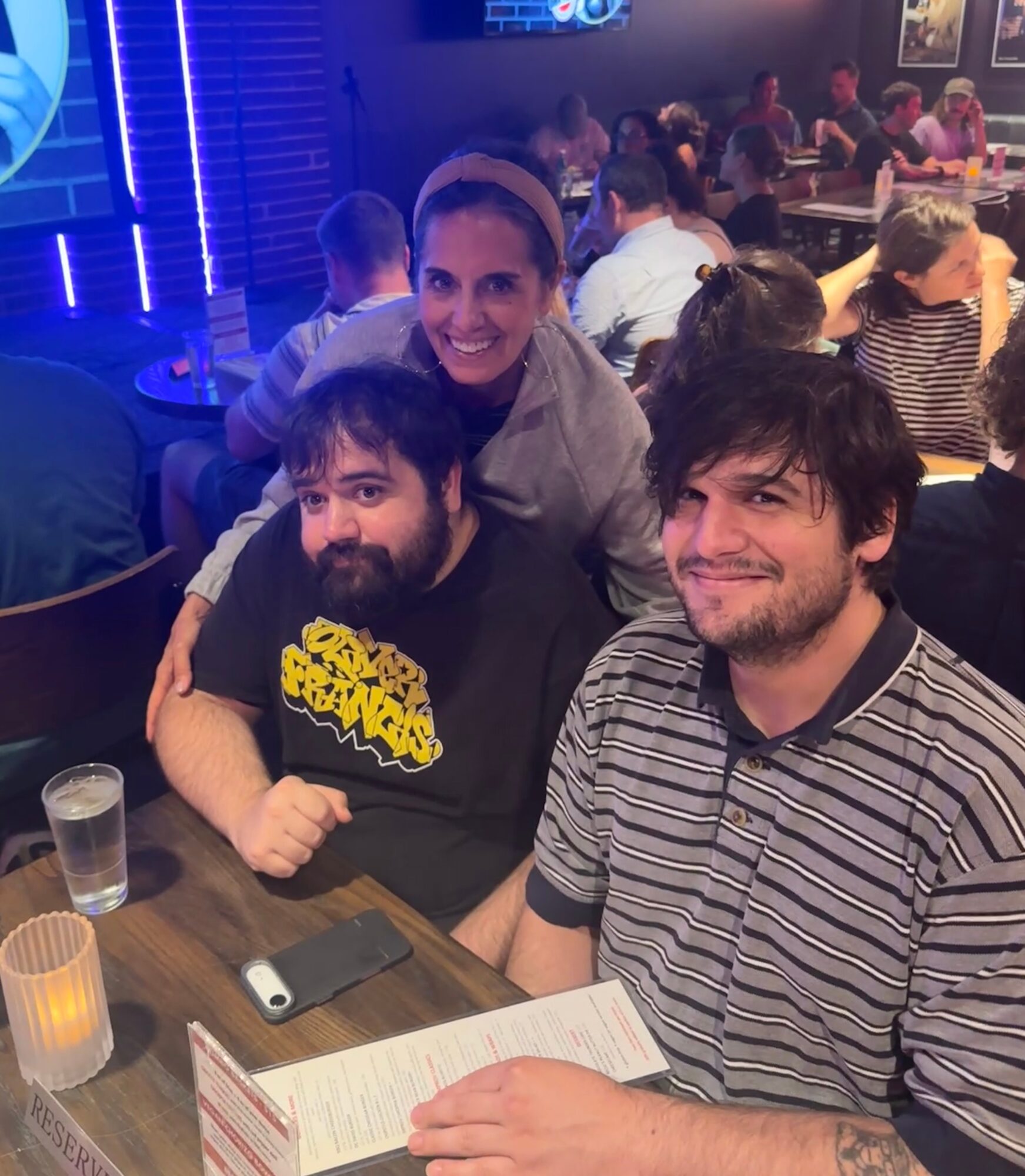 Three people smiling at a table in a crowded indoor setting, with drinks and a menu visible, background with blue lighting.