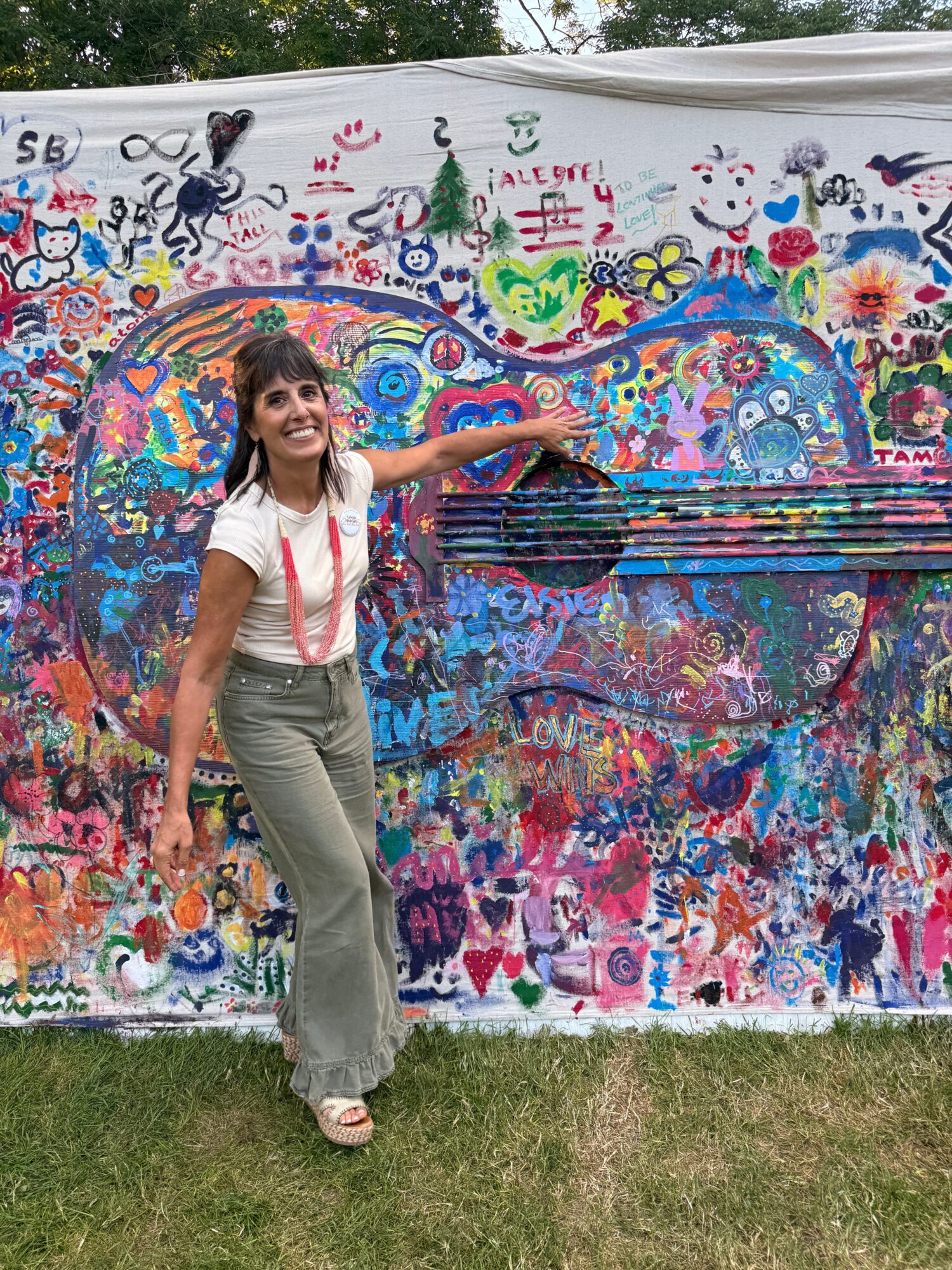 Woman lying on colorful painted mural with various symbols and patterns, outdoors on grass, smiling, wearing gray pants and white top.
