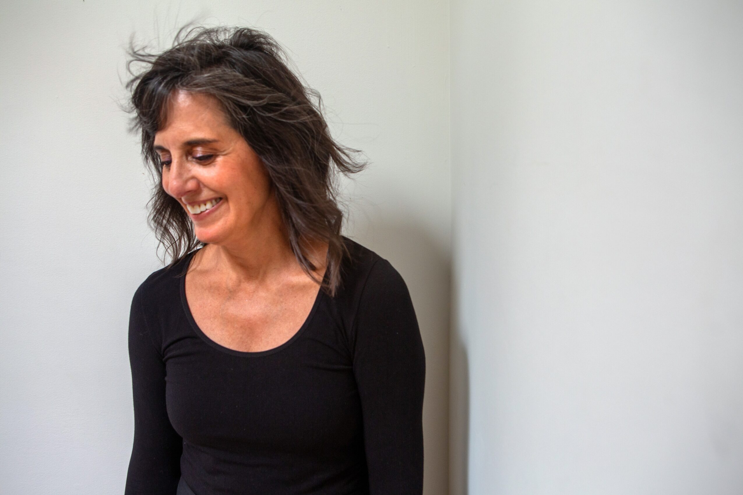 Smiling woman with wavy dark hair wearing a black top, standing against a plain light-colored wall.