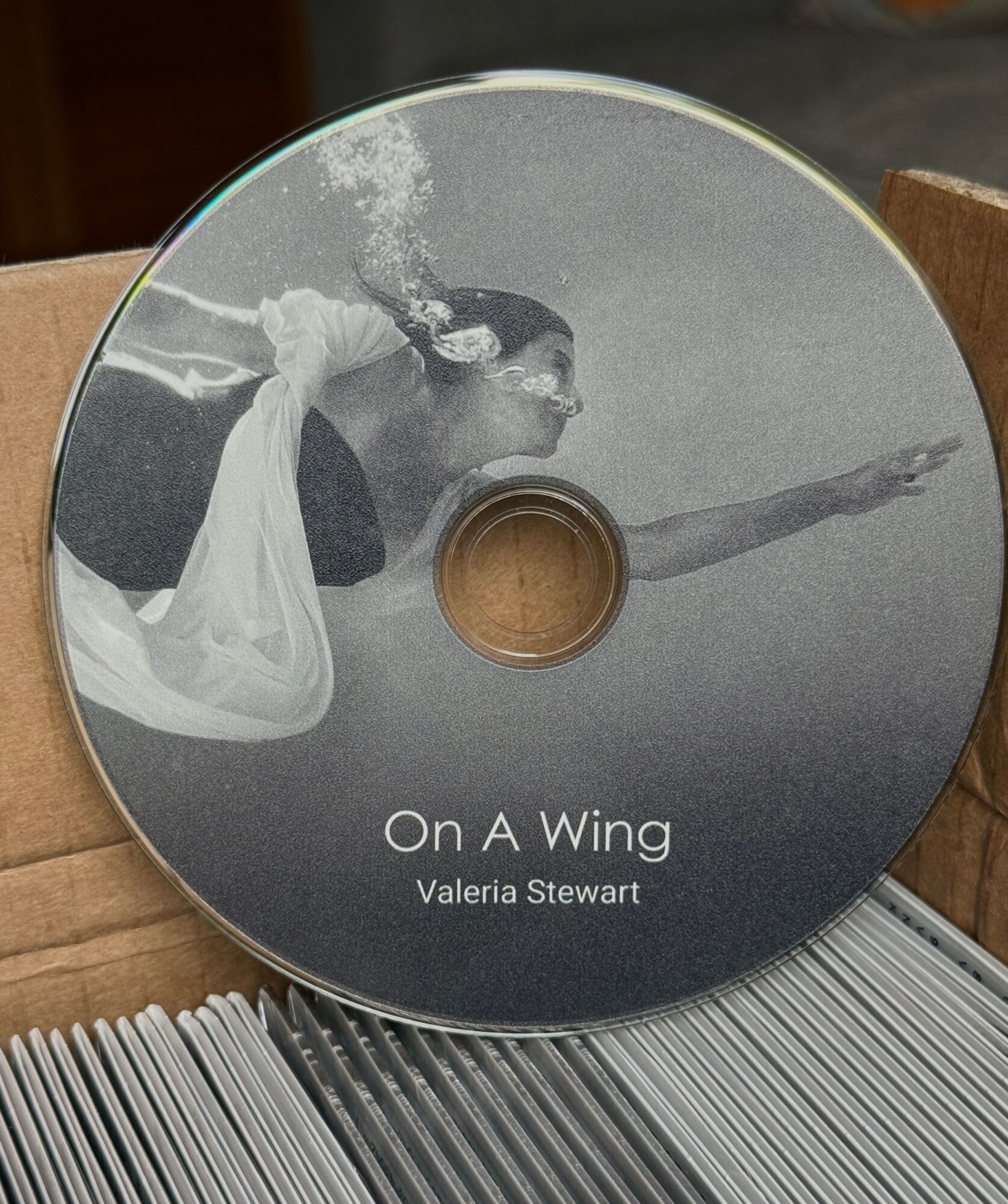 CD with a woman blowing bubbles and reaching forward, titled 'On A Wing' by Valeria Stewart, in a cardboard box.