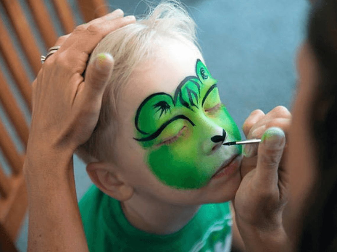 Child's face being painted with a green and black frog design, face paint artist applying details, child's eyes closed, hand on child's head.
