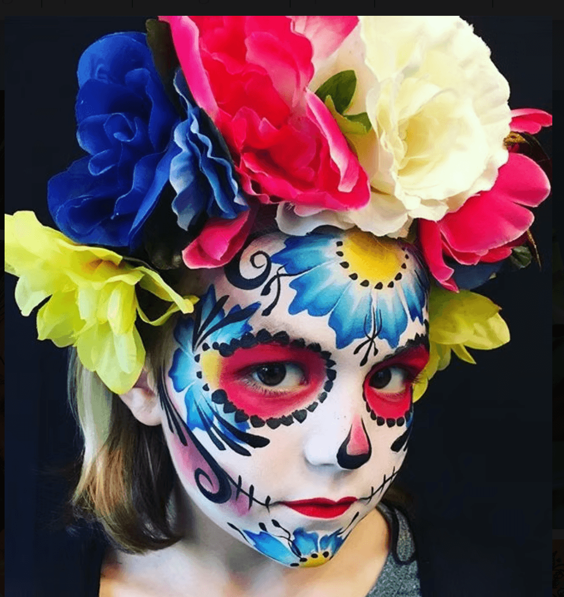 Person with colorful floral headpiece and detailed face paint resembling a sugar skull, with black, white, blue, pink, and yellow colors.