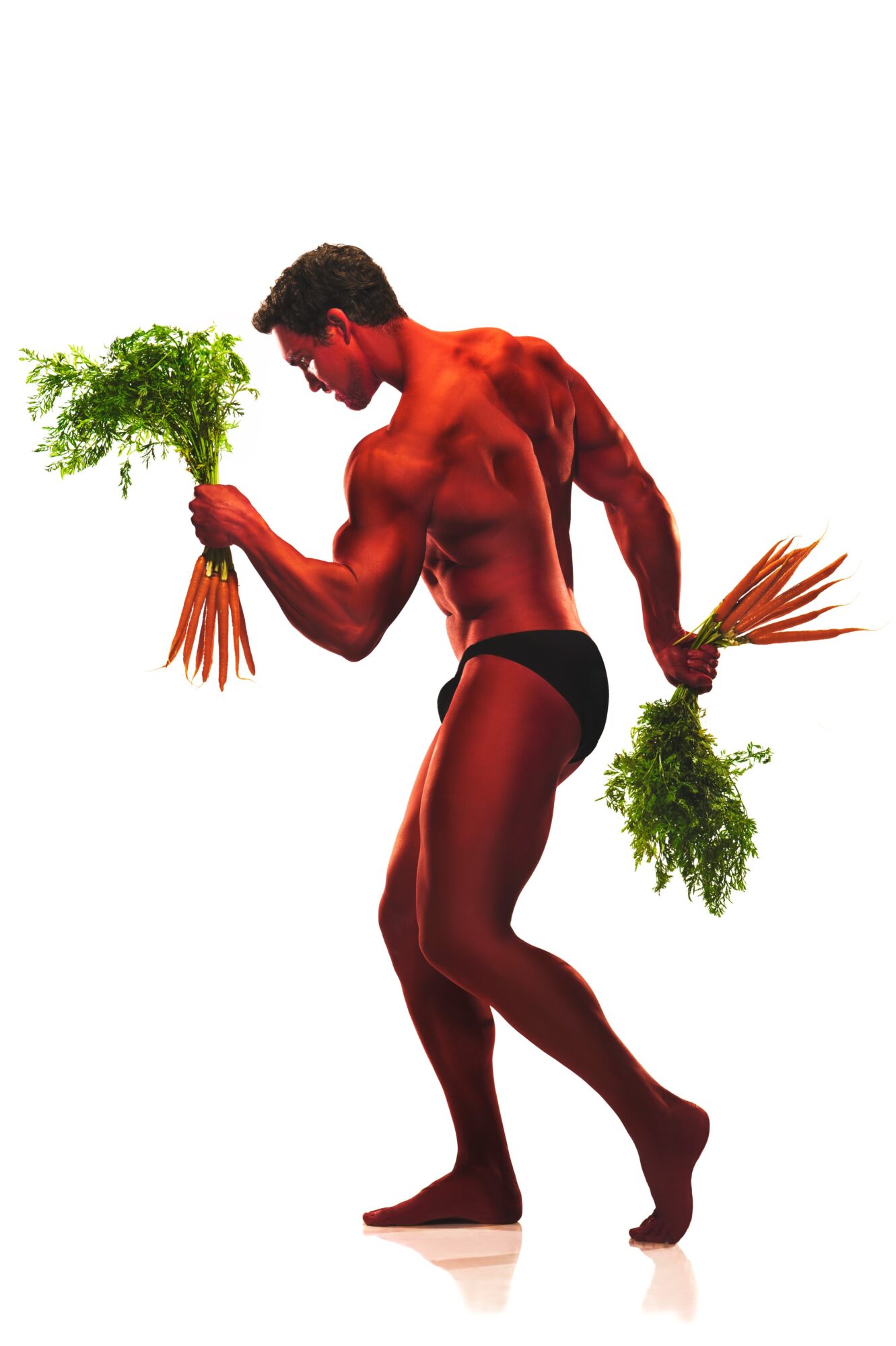 A muscular man with dark hair holds two bunches of green leafy plants, one in each hand, against a white background.
