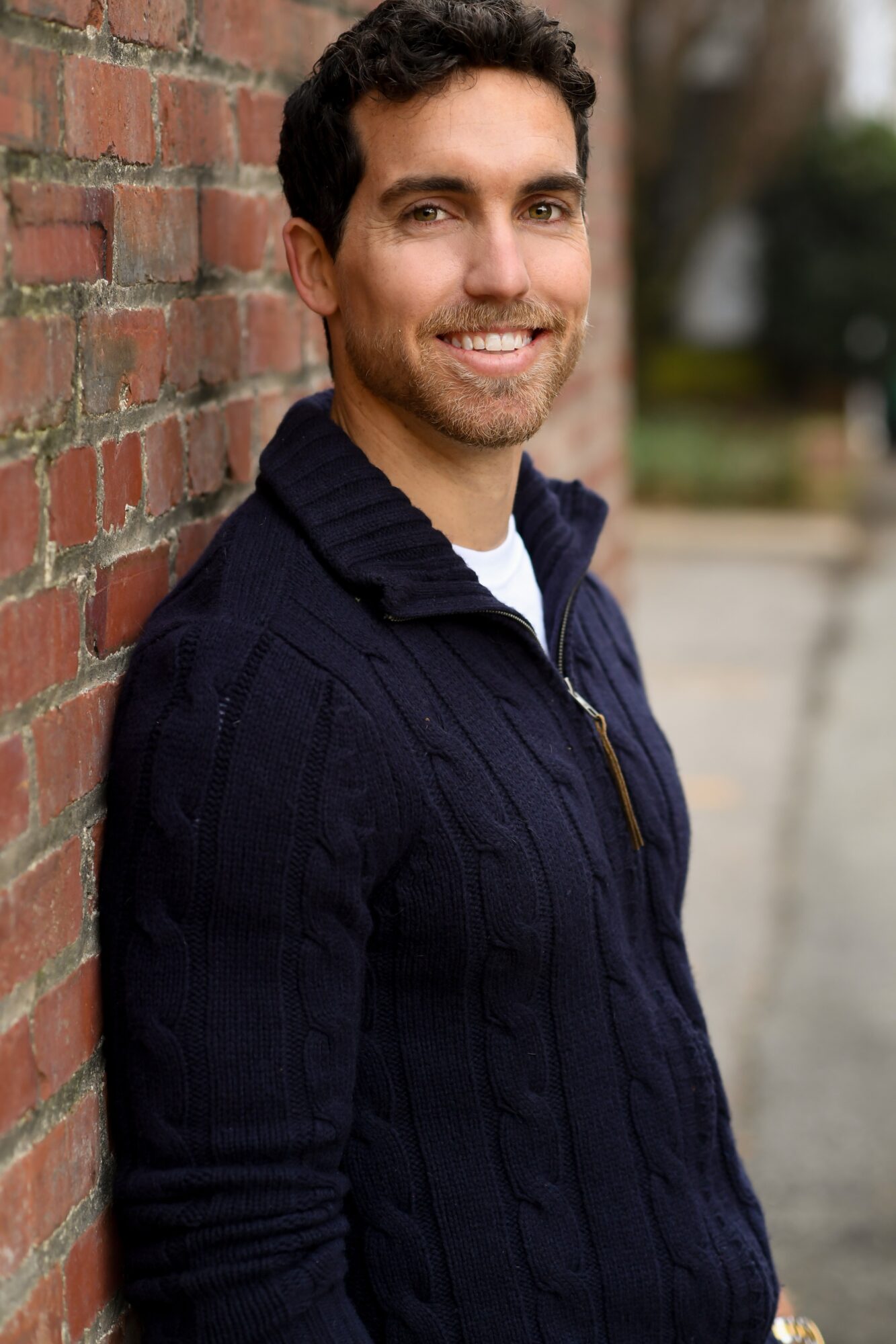 Man with dark curly hair and a beard leaning against a brick wall, smiling outdoors.