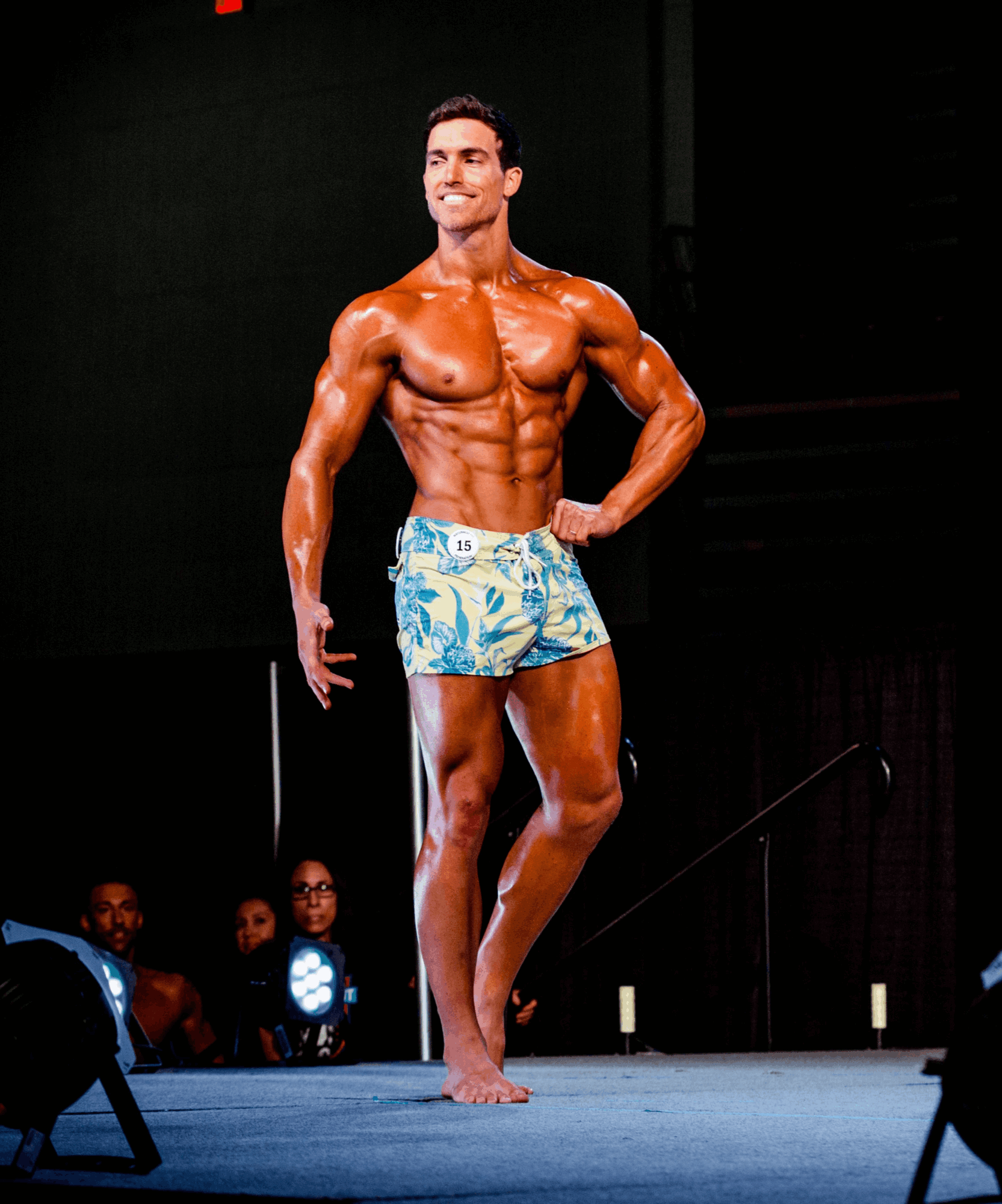 Male bodybuilder on stage in colorful shorts, posing with hand on hip, smiling, with audience in background.