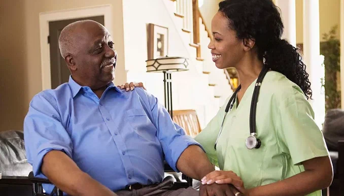 A healthcare worker smiling and talking to an elderly man in a home setting.