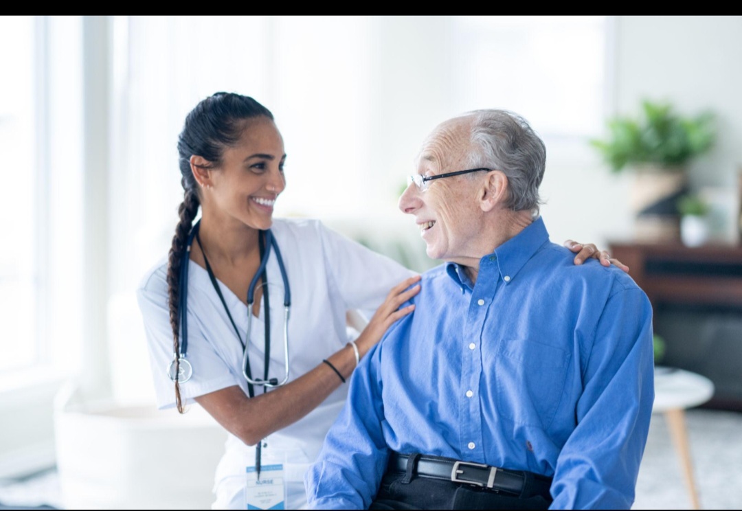 A young female healthcare worker smiling and talking to an elderly man in a bright room.