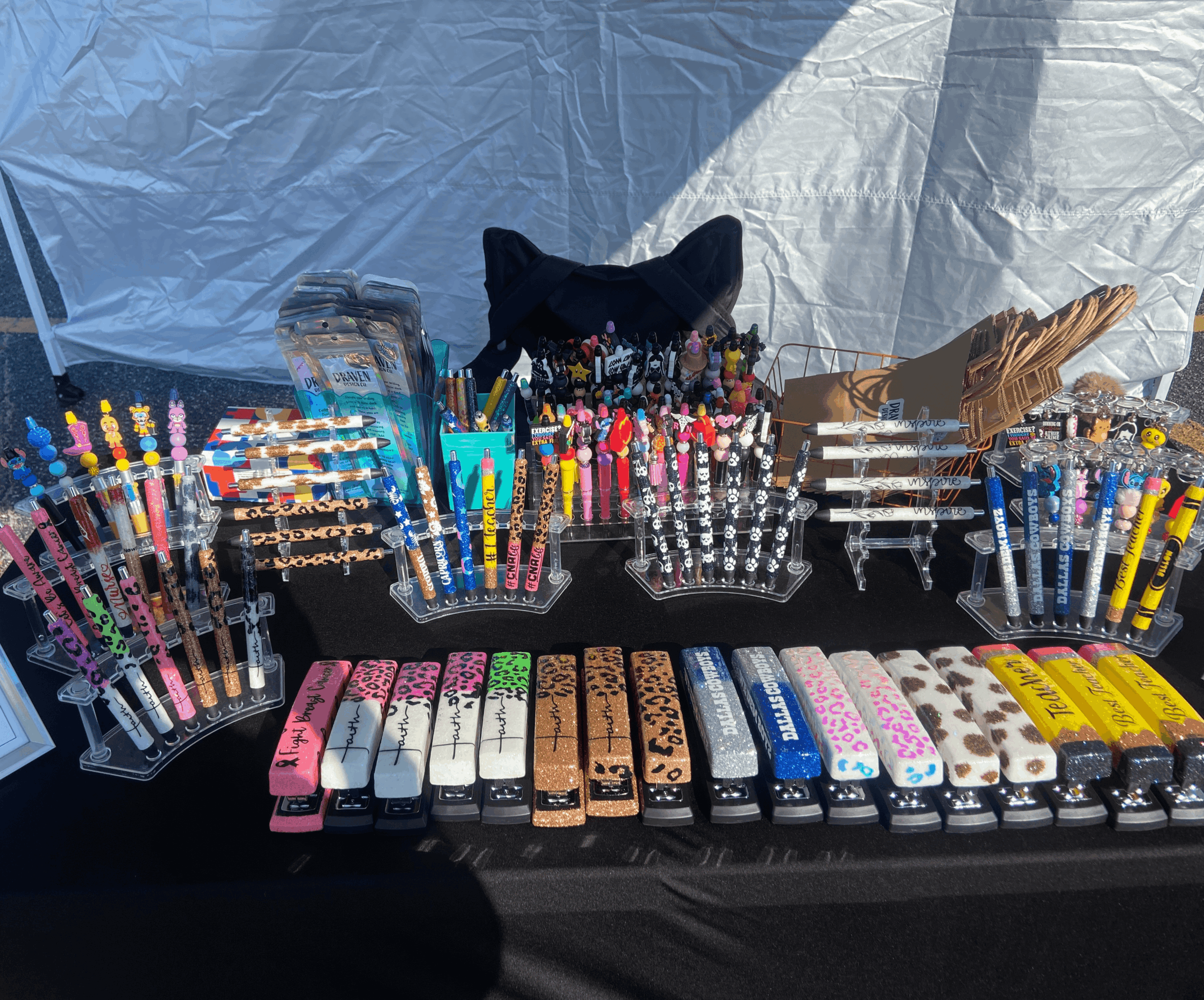 Display of colorful pens, markers, and stationery items on a black table with a white background, including packs of pens and markers.