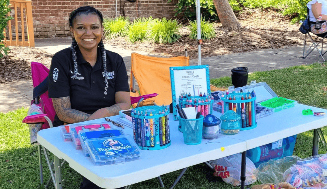 Smiling woman sitting at a table outdoors with various colorful stationery items, trees, and grass in the background.